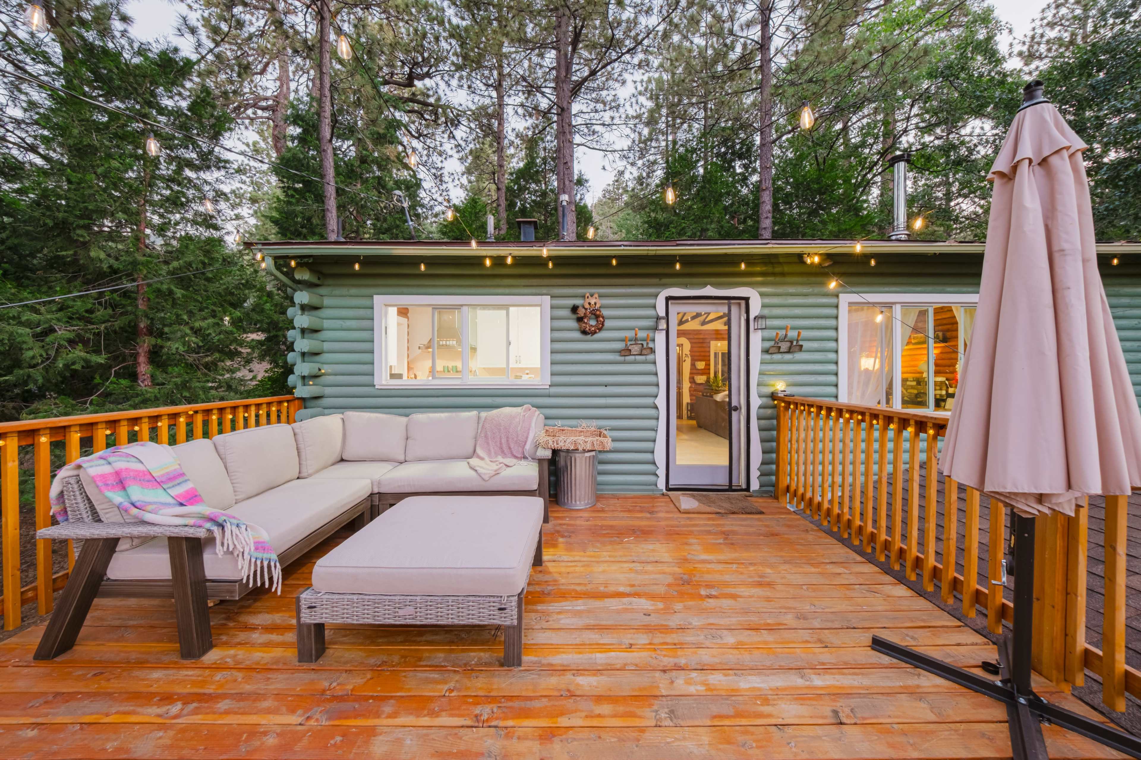 A wooden deck features a sectional sofa and a coffee table, with a green cabin in the background surrounded by tall trees.