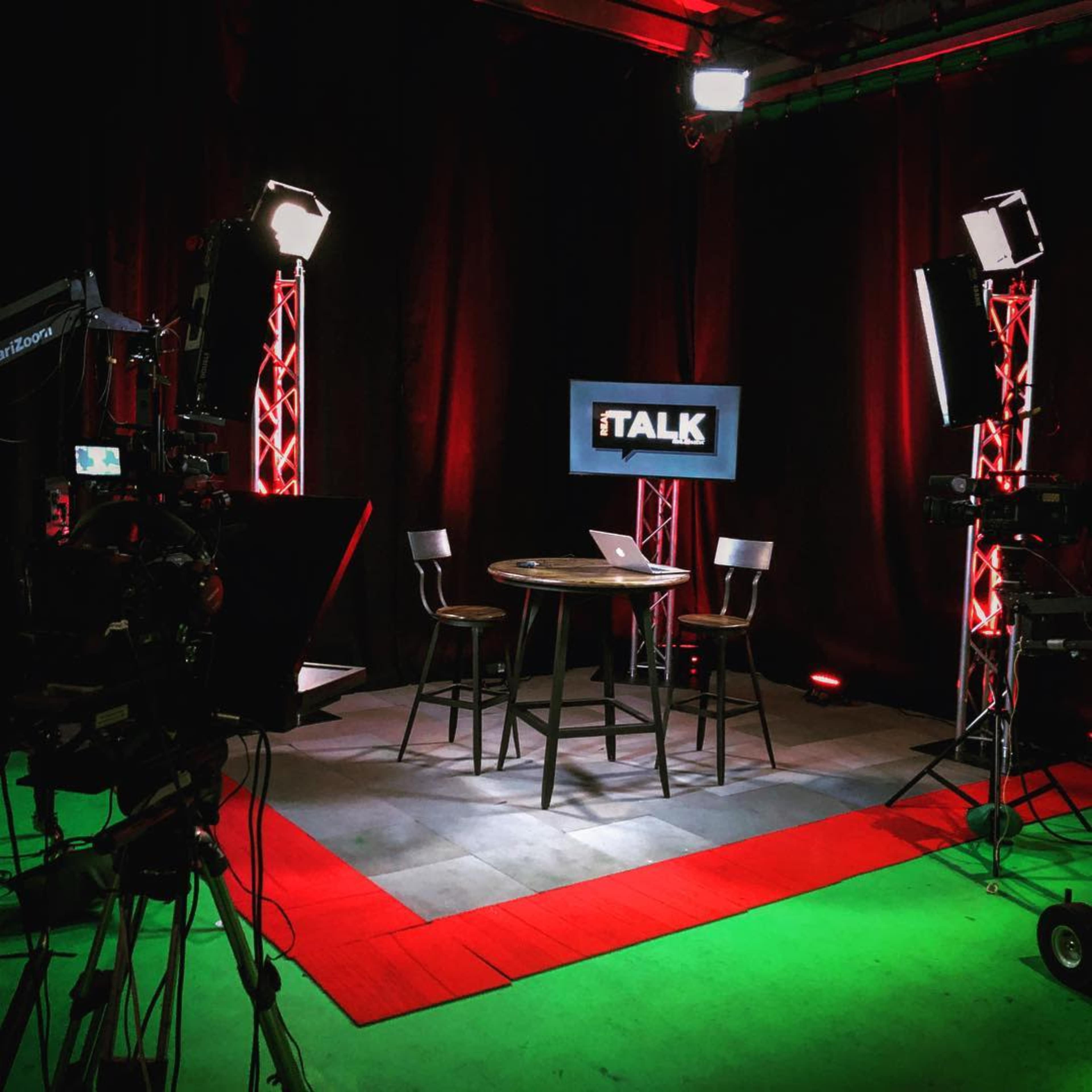 A television studio setup with two empty chairs around a round table, illuminated by multiple lights and backdrop with the logo "iTALK."
