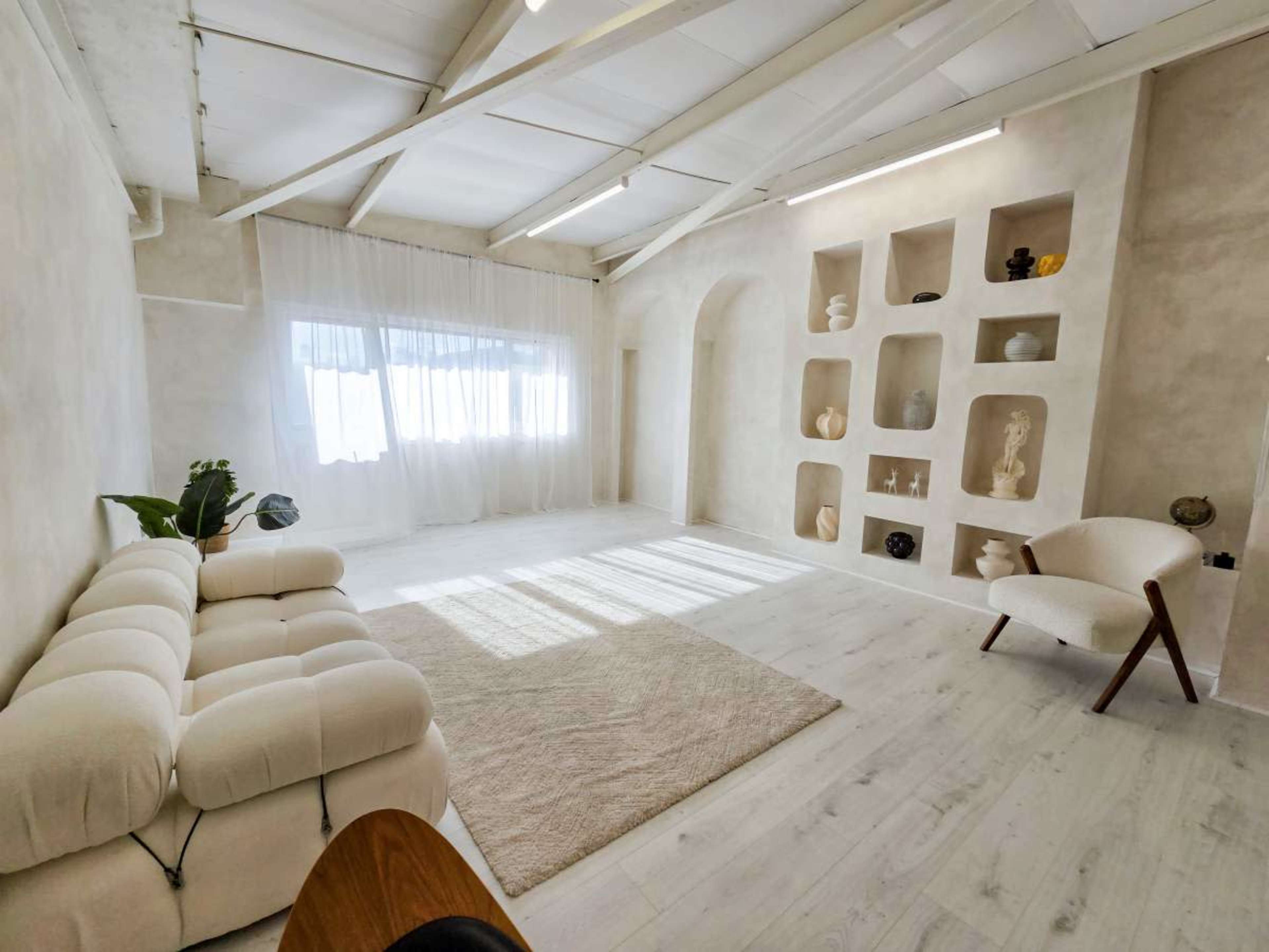 The image shows a spacious, minimalist room featuring a white sofa, a light rug, an armchair, and decorative shelves on the wall.