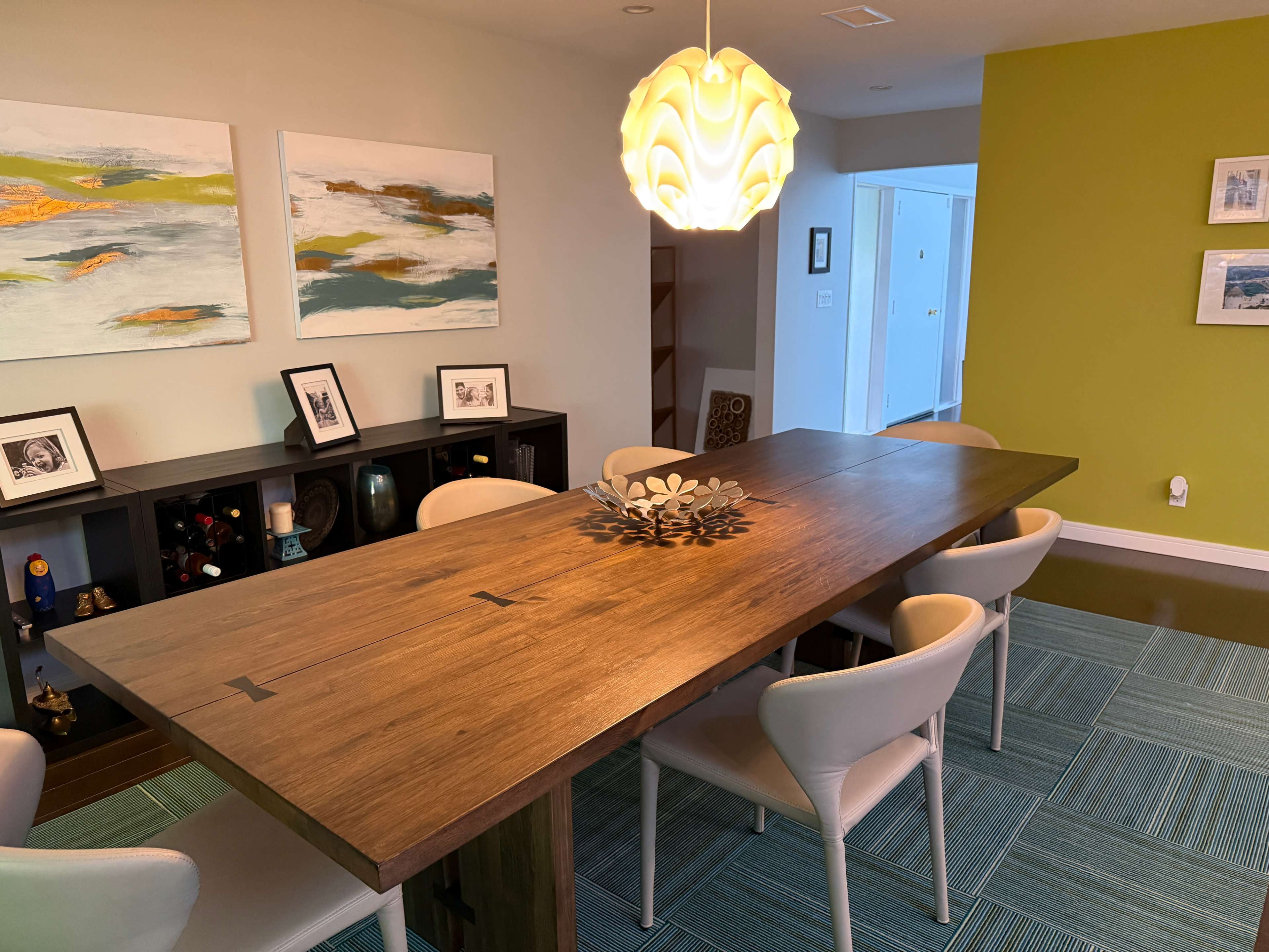 The image shows a modern dining area featuring a long wooden table surrounded by white chairs, with a decorative centerpiece and a pendant light overhead, complemented by colorful artwork on the walls.