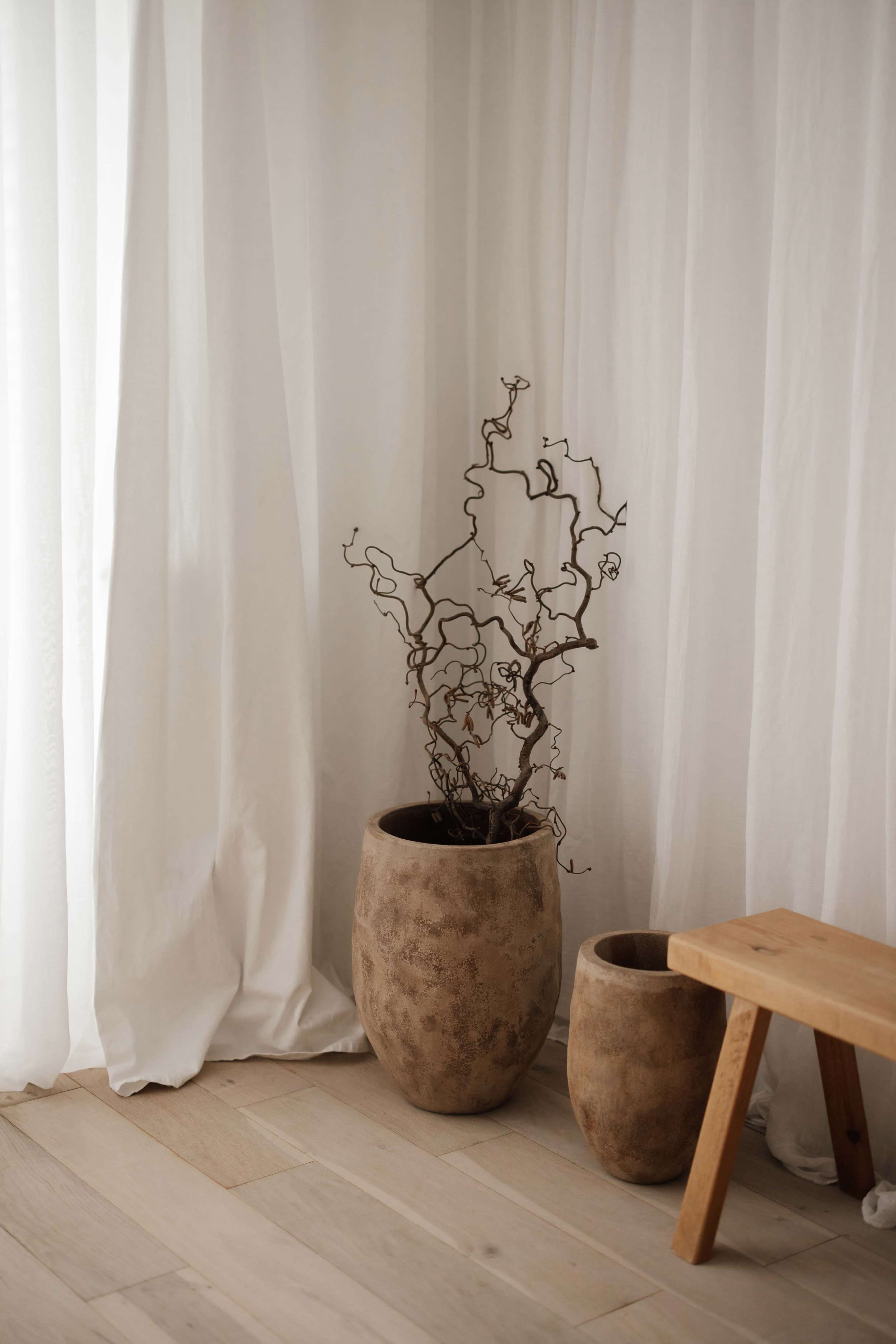A tall, textured vase containing a bare, twisting branch stands next to a smaller vase on a wooden floor, with sheer white curtains in the background.
