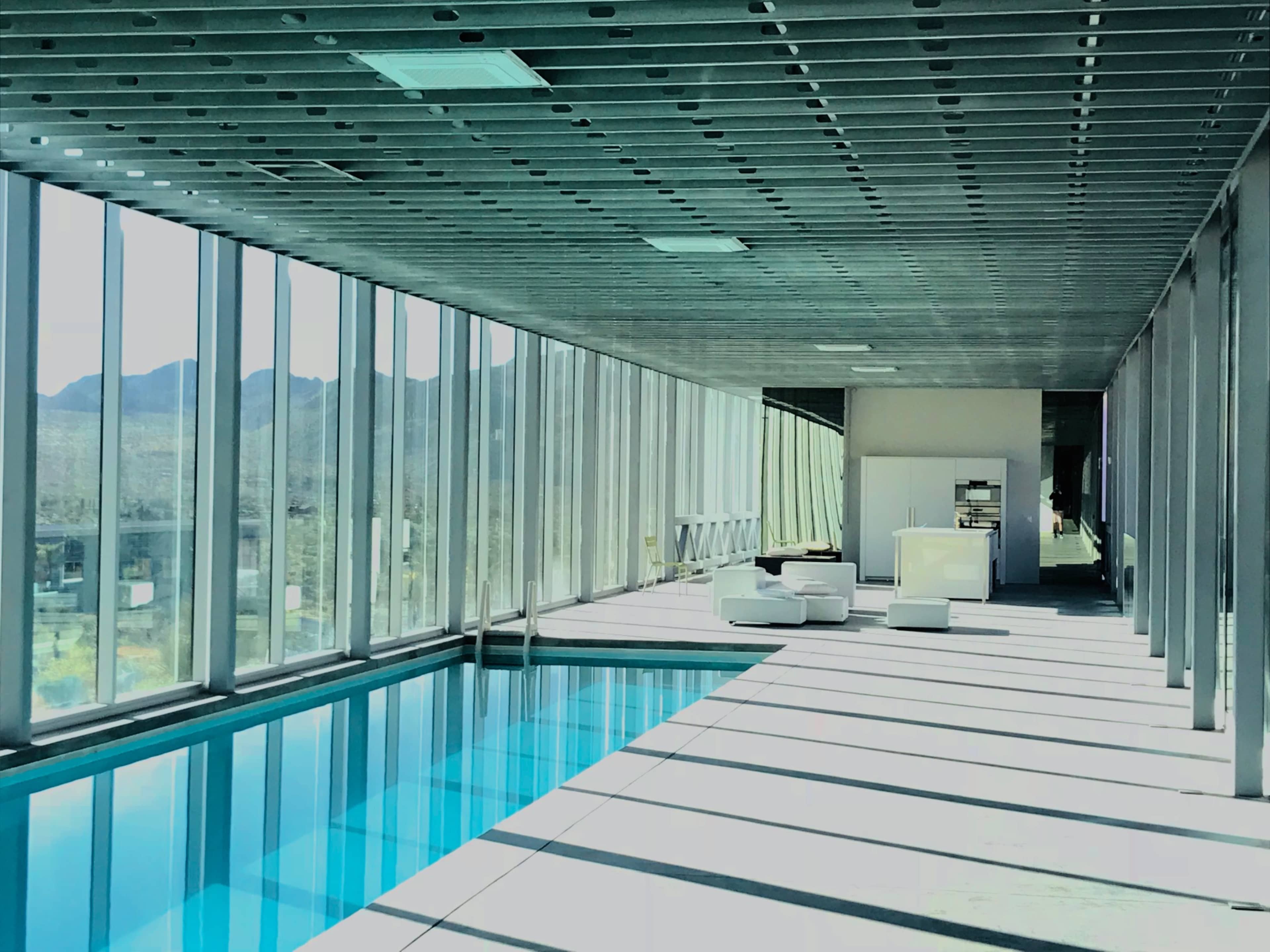 A modern interior space with a floor-to-ceiling glass wall, a swimming pool on one side, and minimalist furniture.