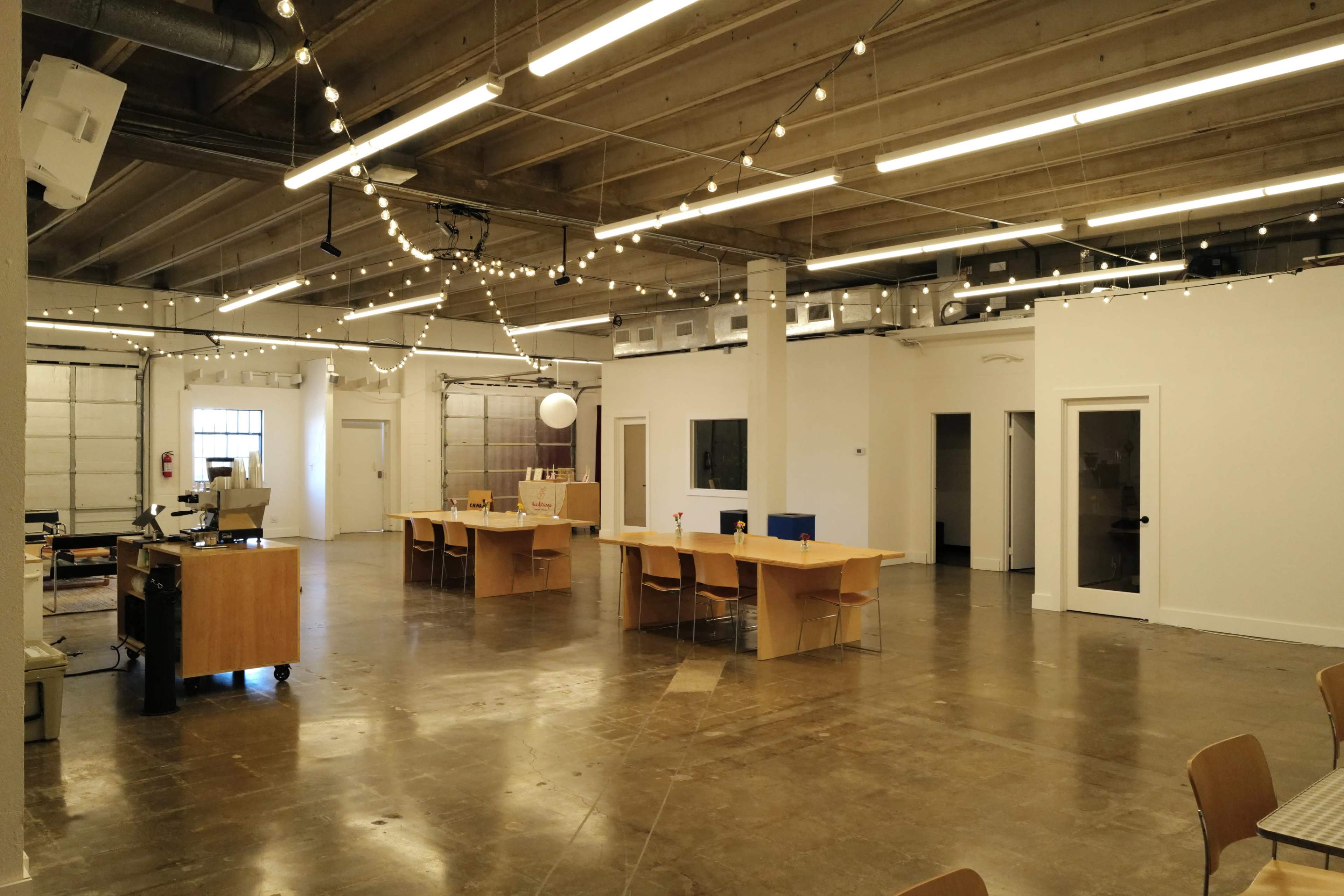 The image shows a spacious, modern office environment with several wooden tables and chairs, string lights overhead, and white walls.