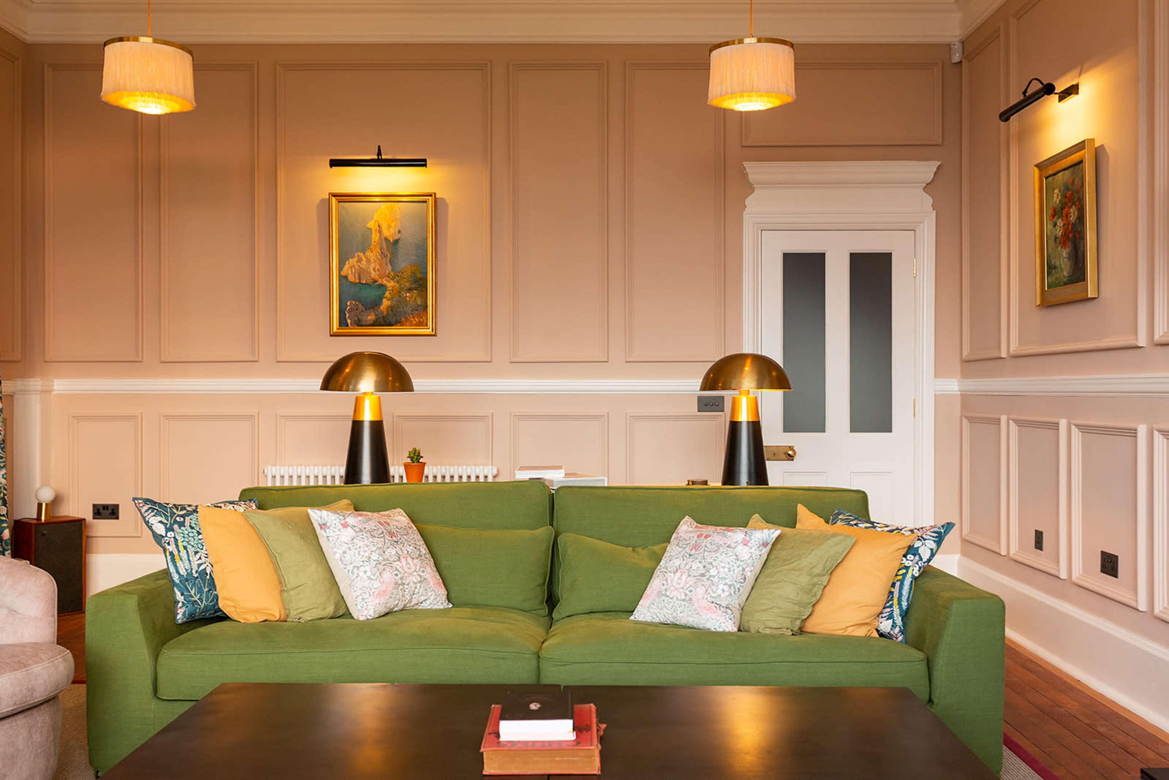 A green sofa with patterned cushions in a room featuring beige paneling, gold lamps, and wall art.