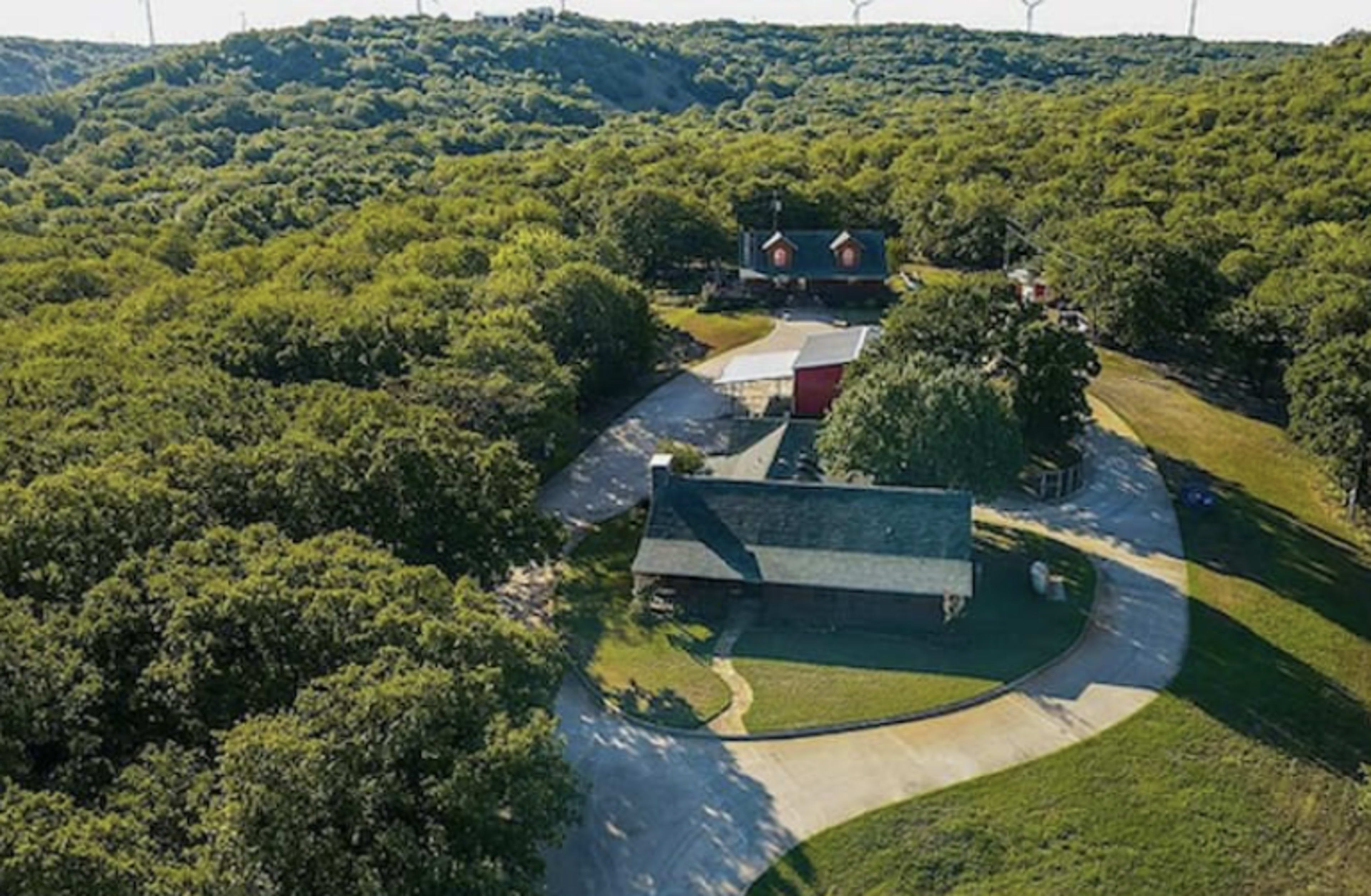 Aerial view of a residential property surrounded by dense green trees, featuring multiple buildings and a winding driveway.