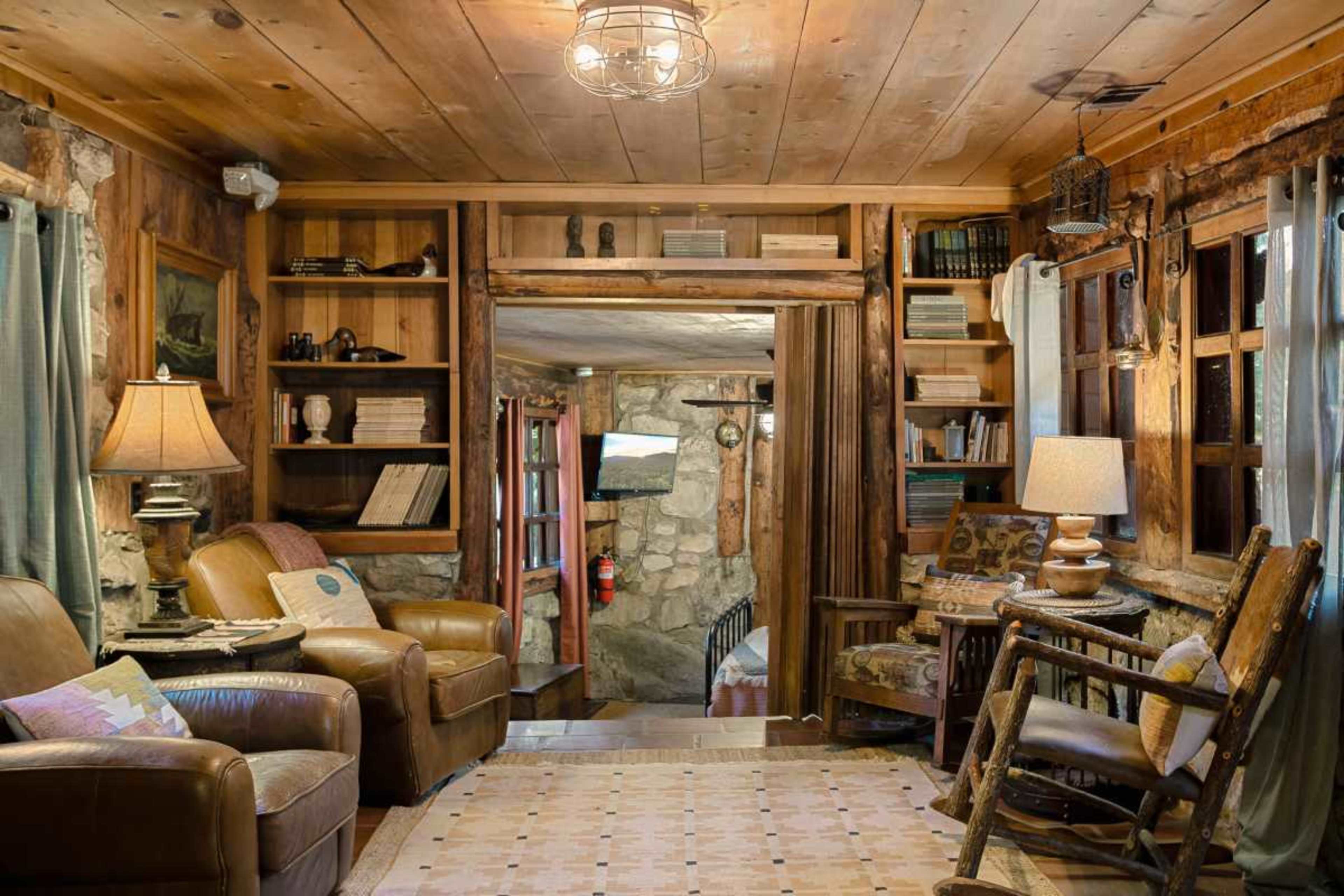 The image shows a cozy living room with wooden walls, two leather armchairs, a lamp, bookshelves filled with books, and a view into a stone-walled corridor.