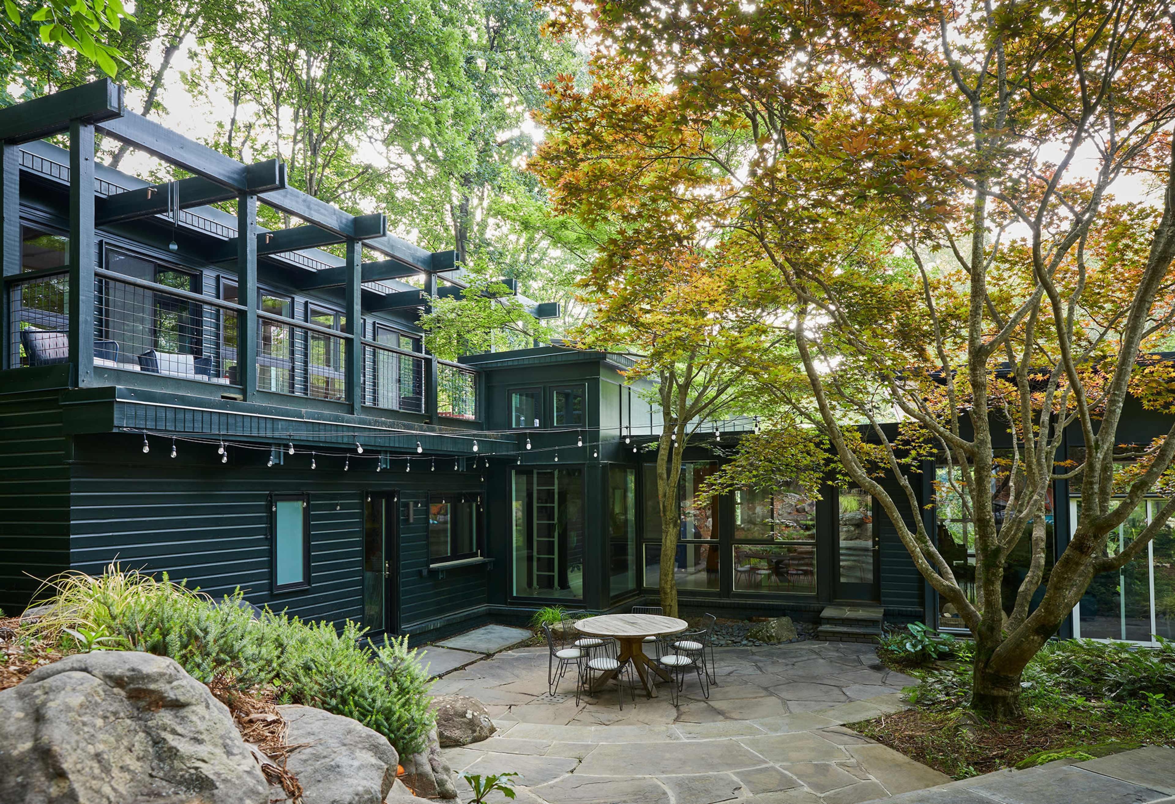 The image shows a modern house with dark exterior walls, surrounded by lush greenery and a stone patio with a circular table.