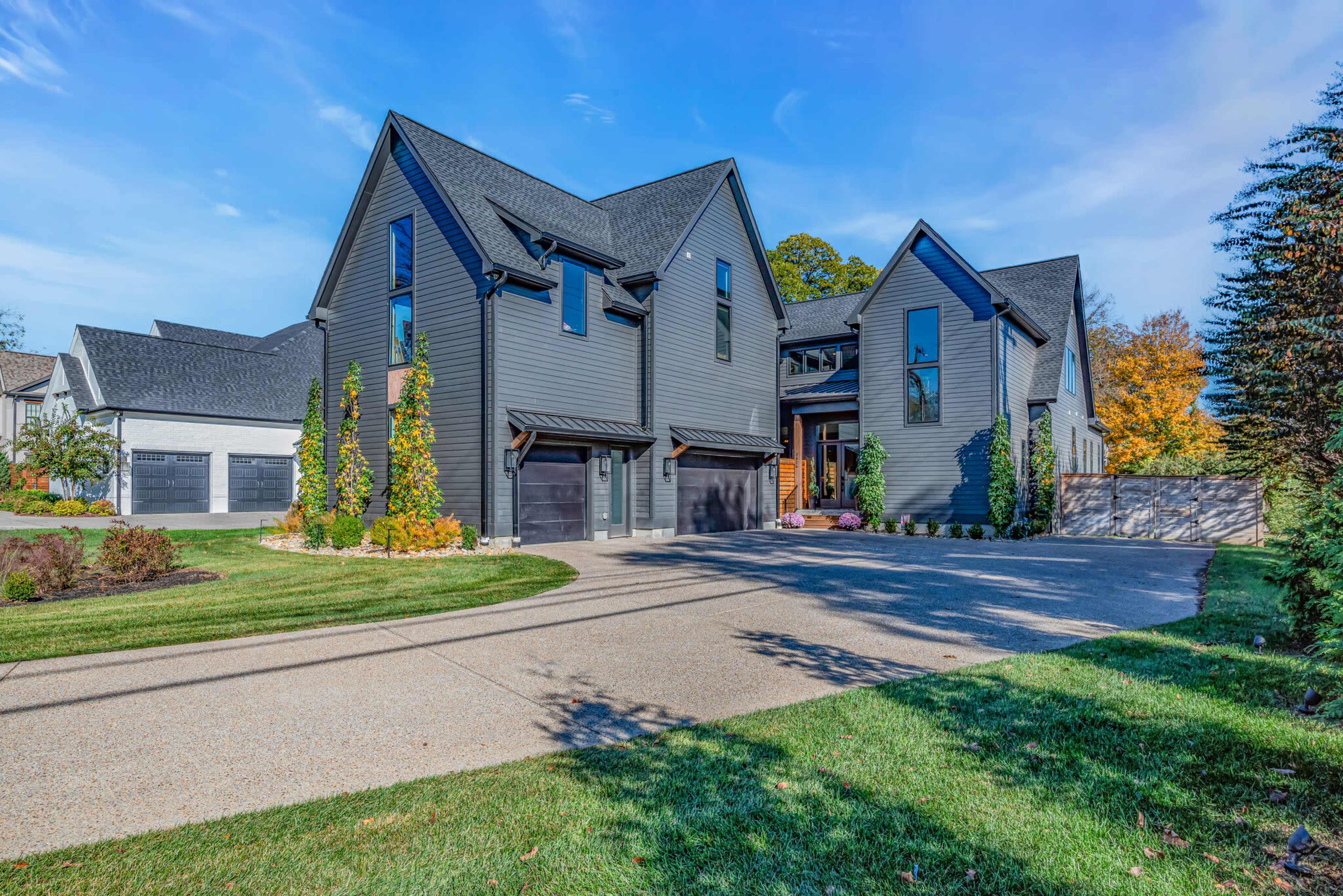 The image shows a modern, two-story house with a dark exterior, surrounded by well-maintained landscaping and a circular driveway.