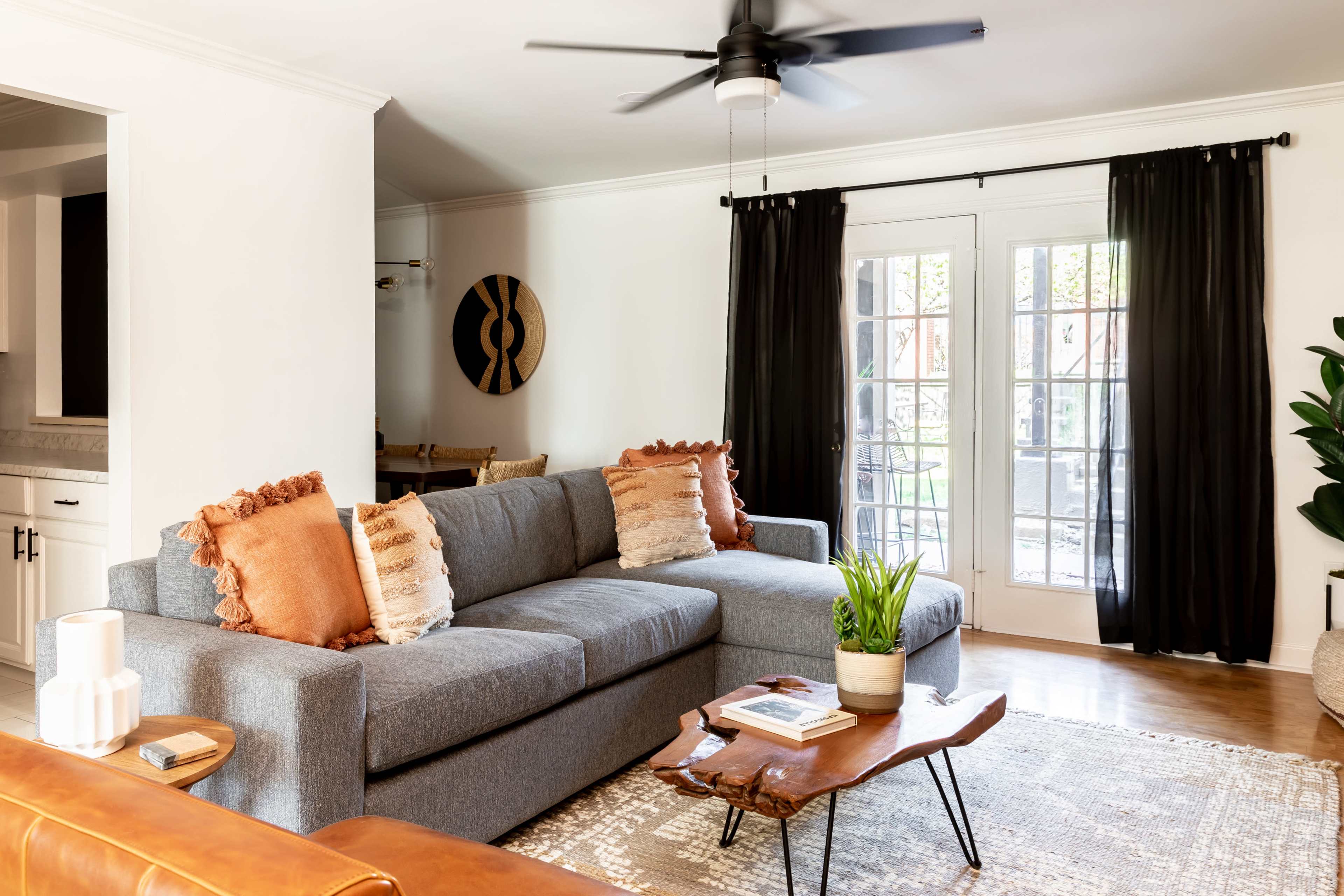 A modern living room features a gray sectional sofa with decorative pillows, a wooden coffee table, and large windows covered by black curtains.