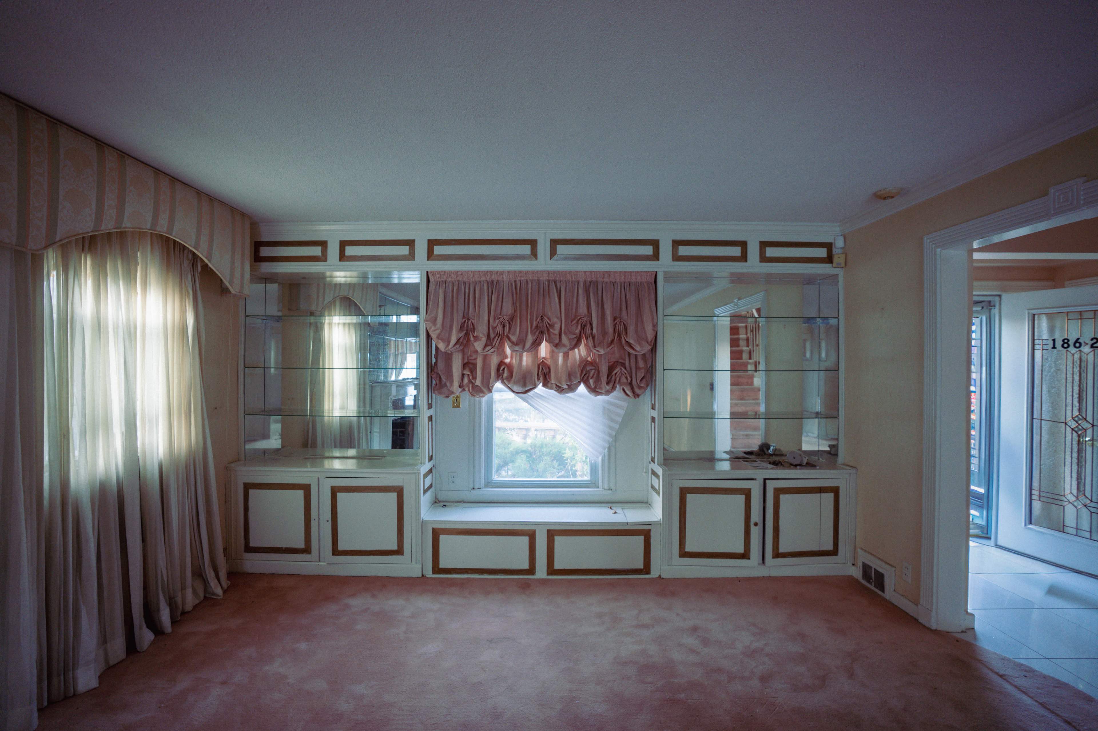 The image shows a softly lit living room featuring a large window with a decorative above, built-in shelves on either side, and light-colored carpeting.
