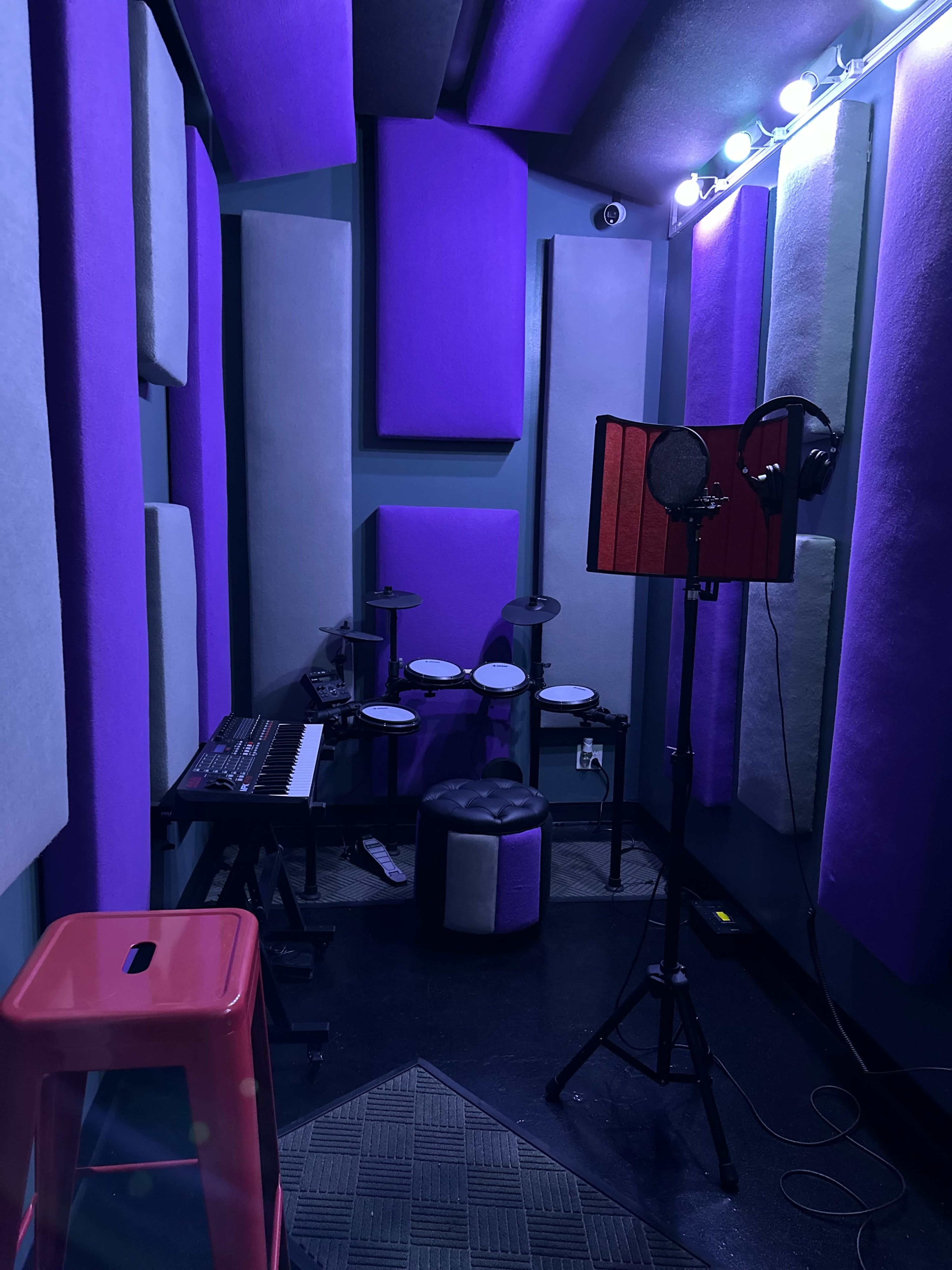 The image shows a small soundproof recording studio with purple acoustic panels, a keyboard, electronic drums, and a microphone setup.