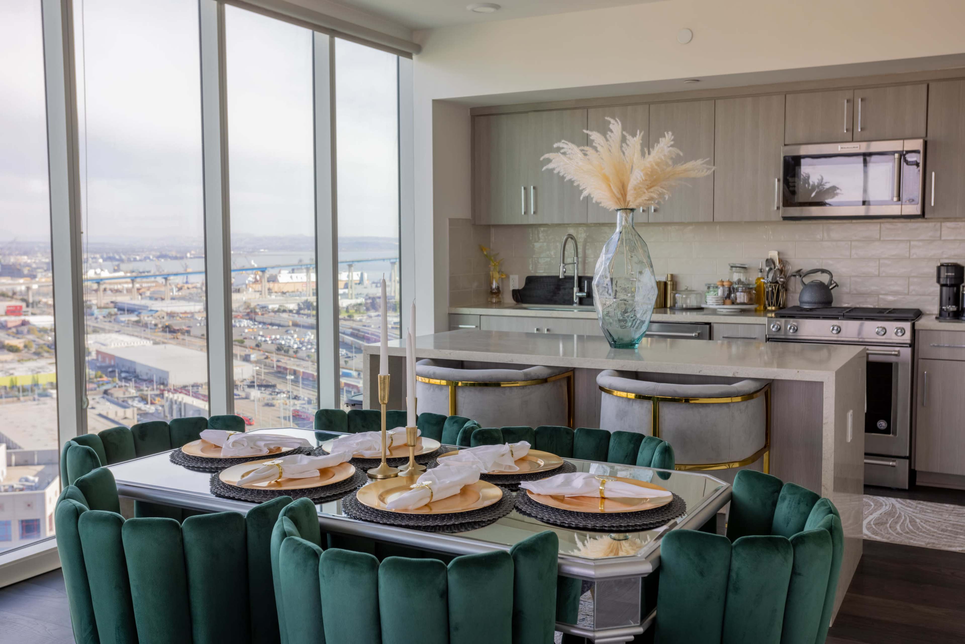 A modern dining area features a circular table set with plates and utensils, surrounded by plush green chairs, with a large window overlooking a cityscape.