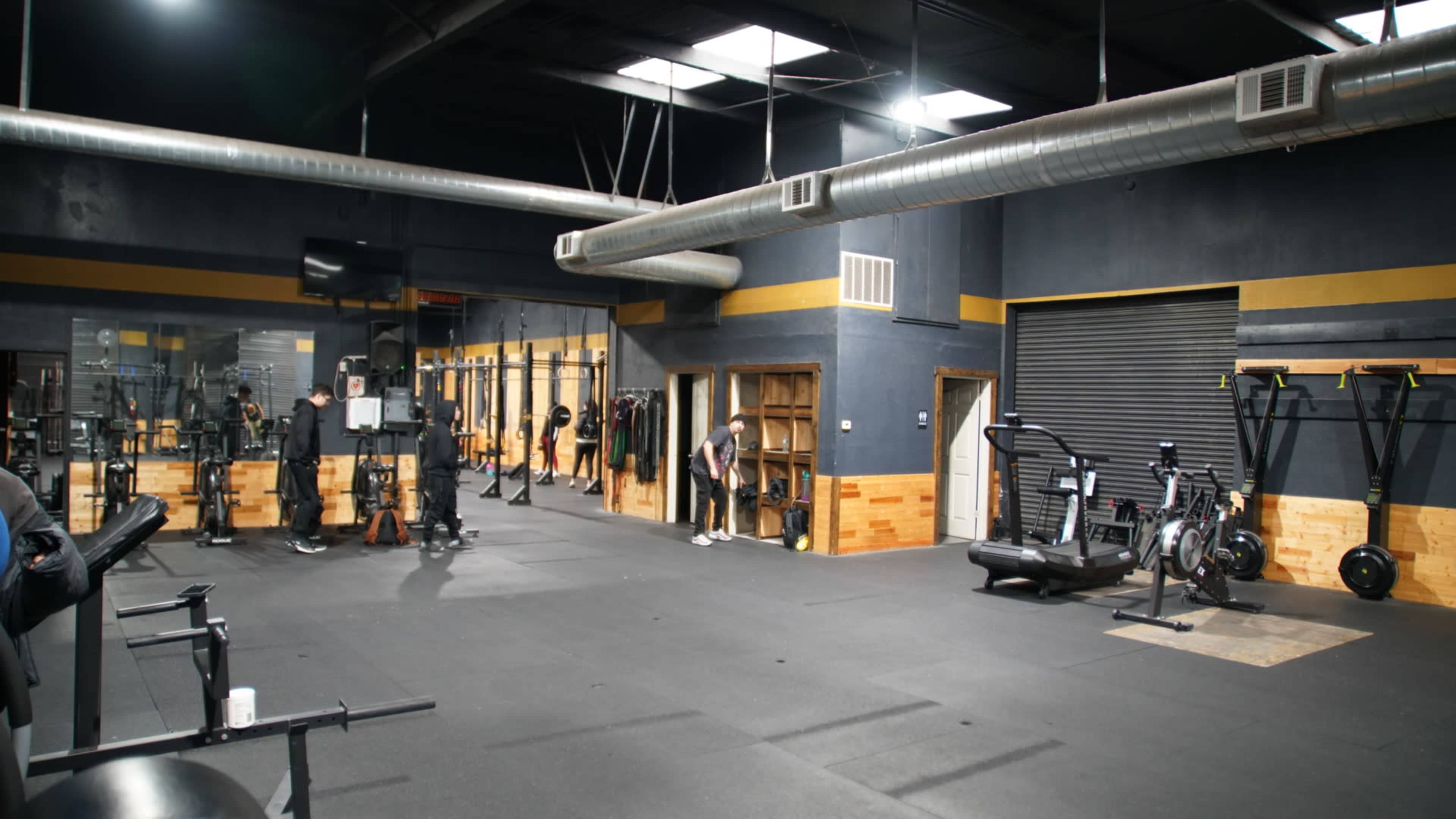 An indoor gym space with exercise equipment, including treadmills, weight racks, and a rowing machine, under bright industrial lighting.