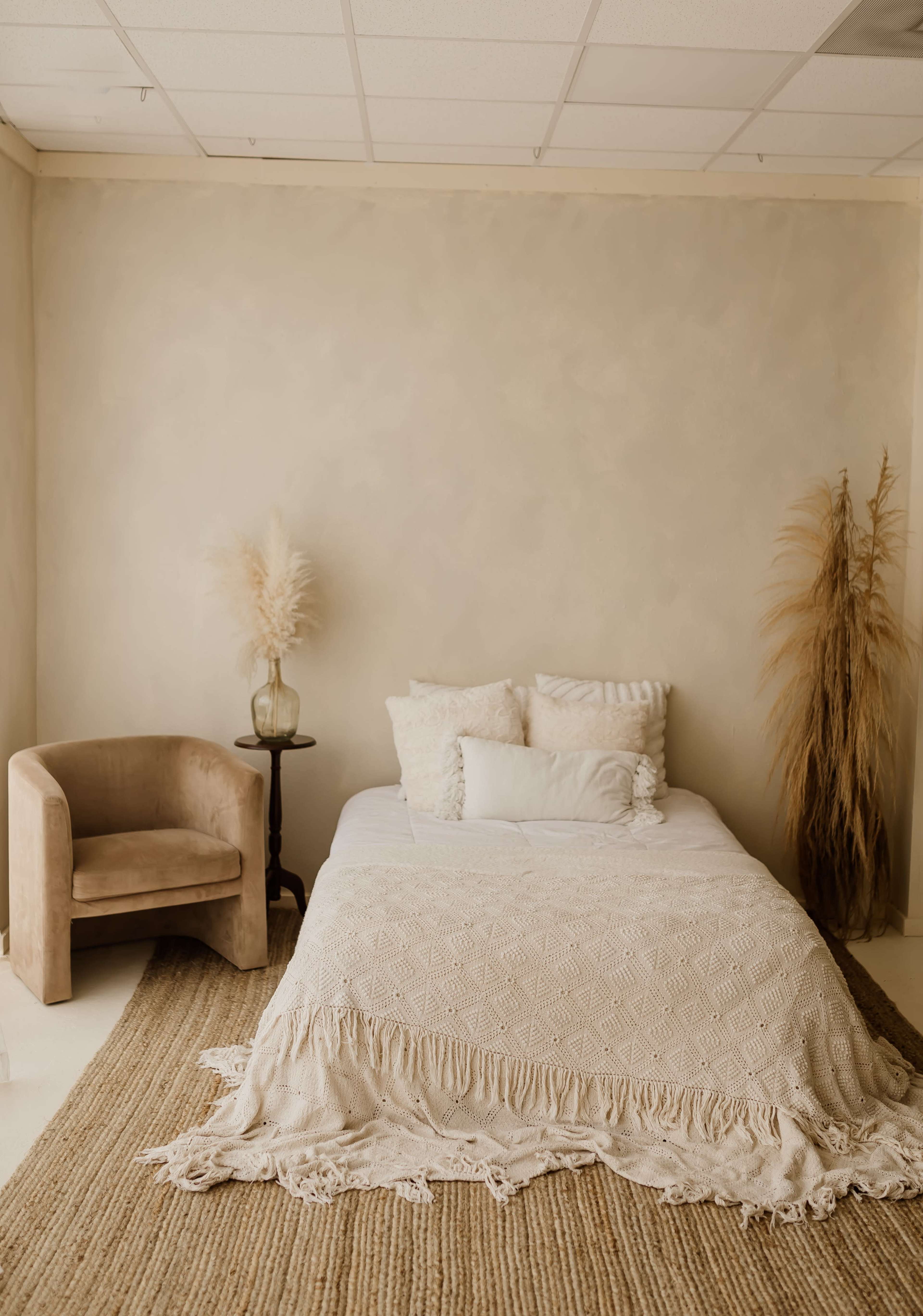 A minimalistic bedroom featuring a bed with a textured blanket, a chair, and decorative dry plants against a neutral wall.