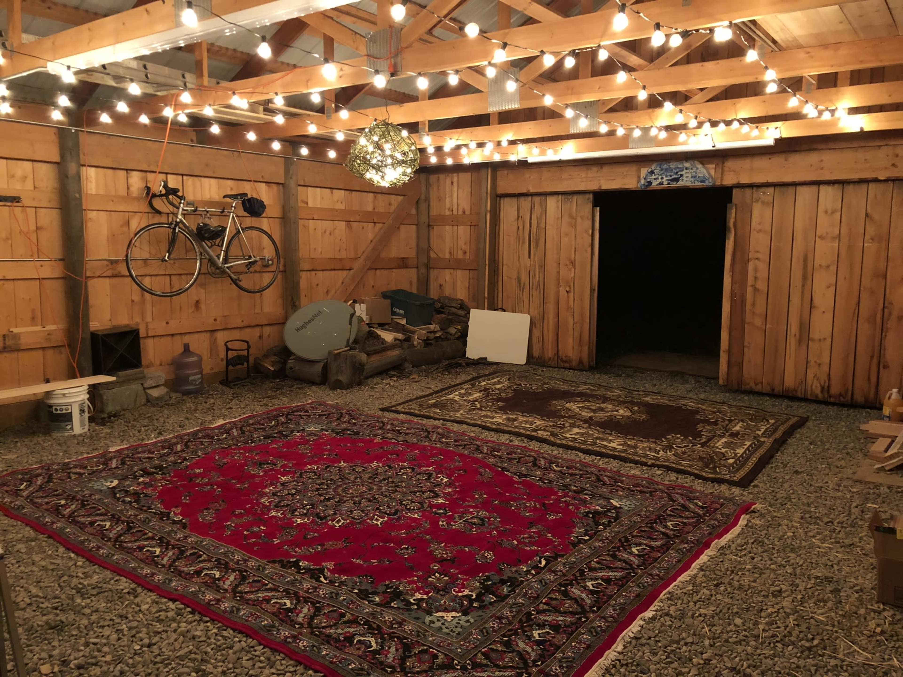 The image shows a rustic barn interior with wooden walls, string lights overhead, and two large rugs on a gravel floor.