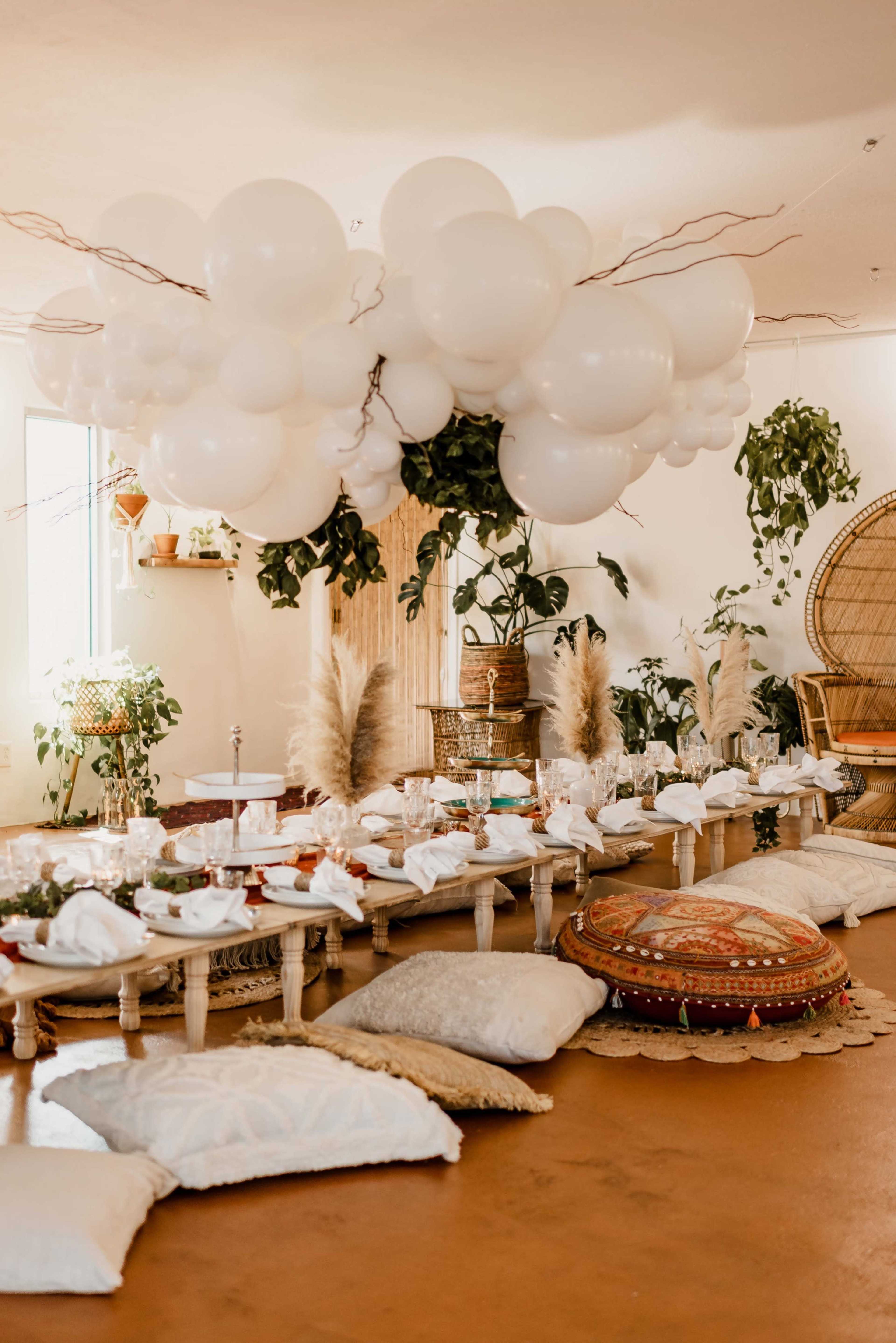 A spacious room features a long table set with plates and napkins, surrounded by decorative cushions and topped with a cloud-like arrangement of white balloons and greenery.