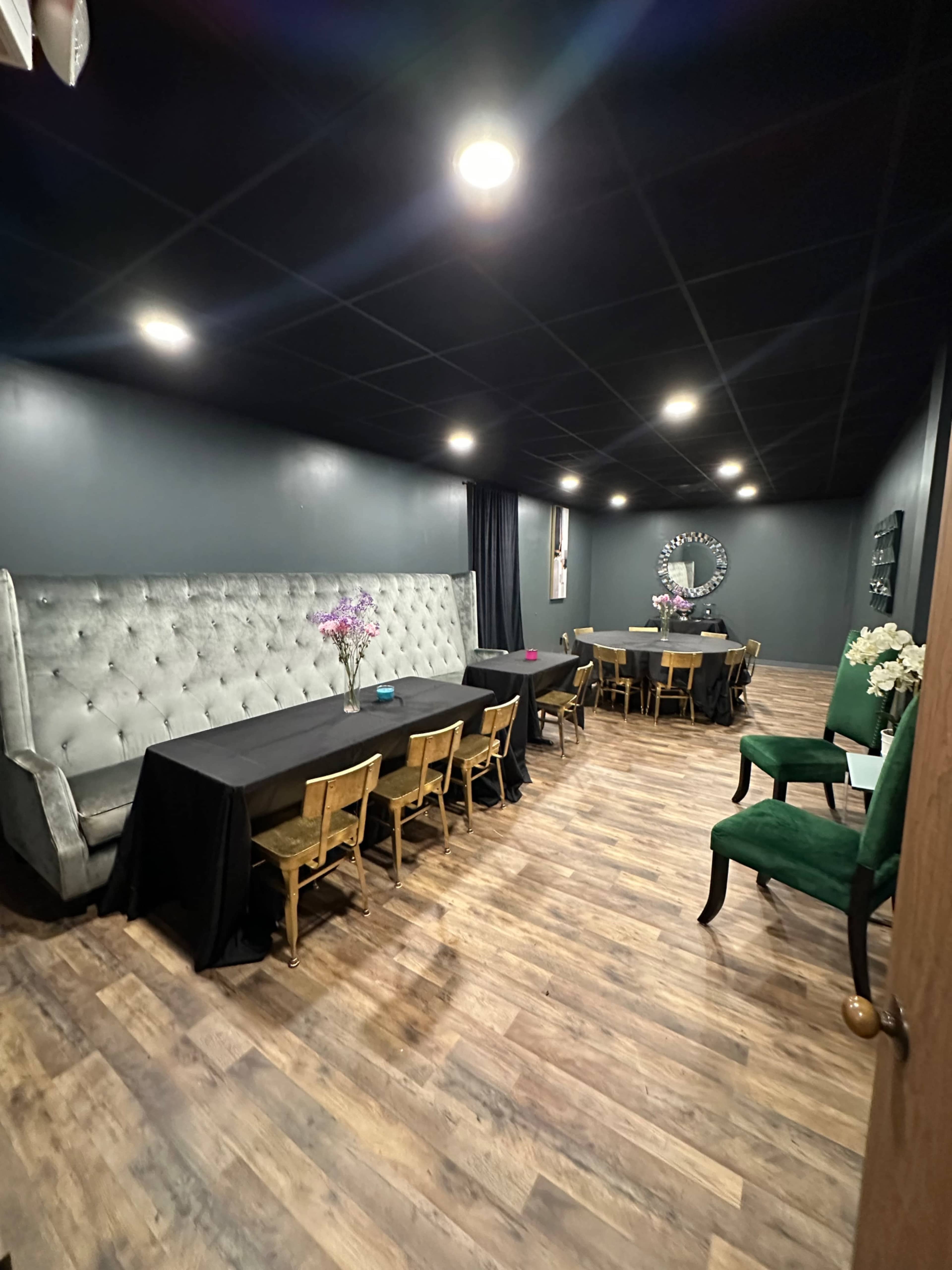 The image shows a dimly lit room with gray walls, featuring a mix of tables and chairs, including a large upholstered seating area and several smaller dining tables arranged throughout the space.