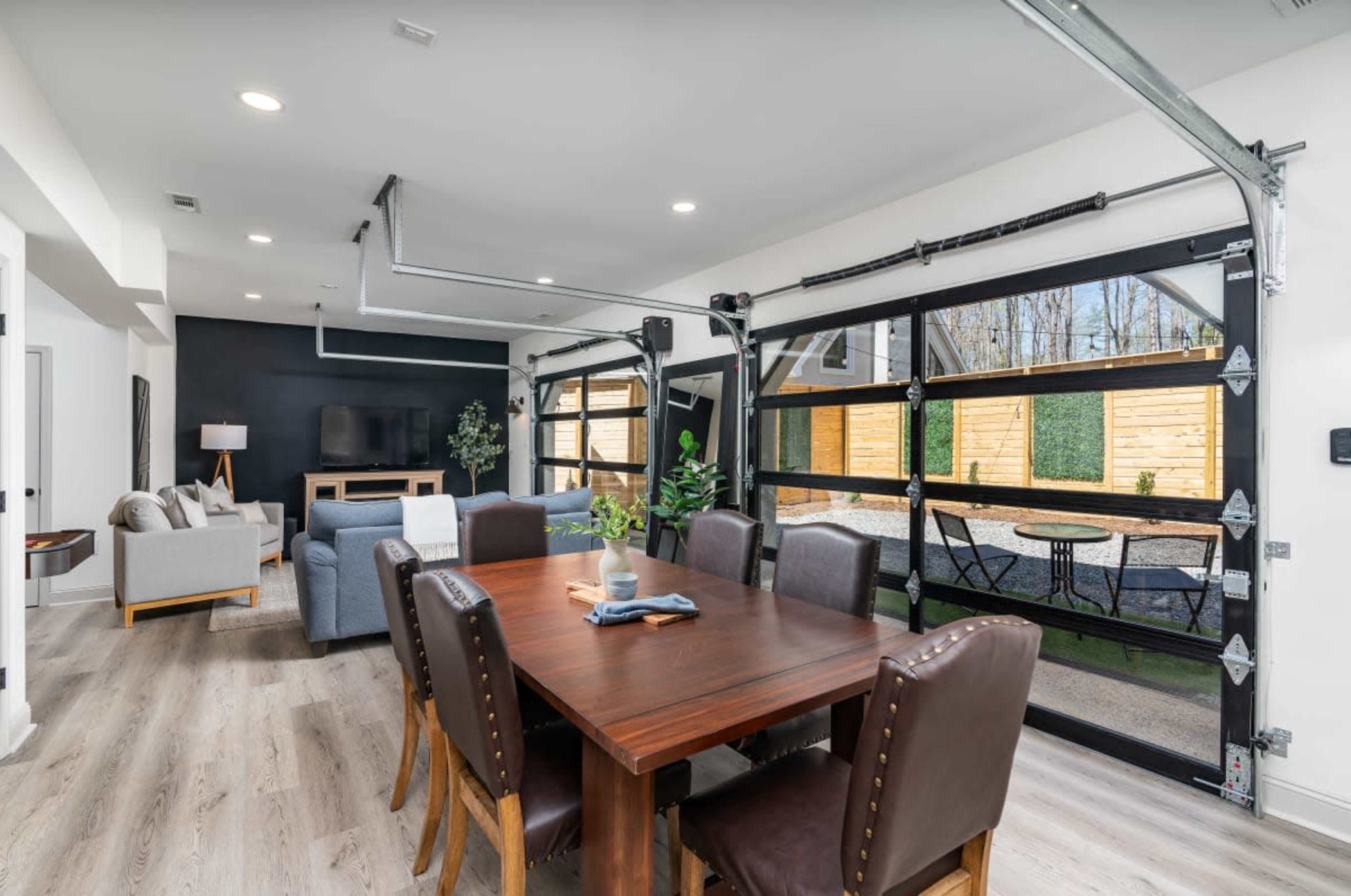 A modern open-concept dining area features a wooden table surrounded by dark leather chairs, with large garage doors opening to a backyard space.