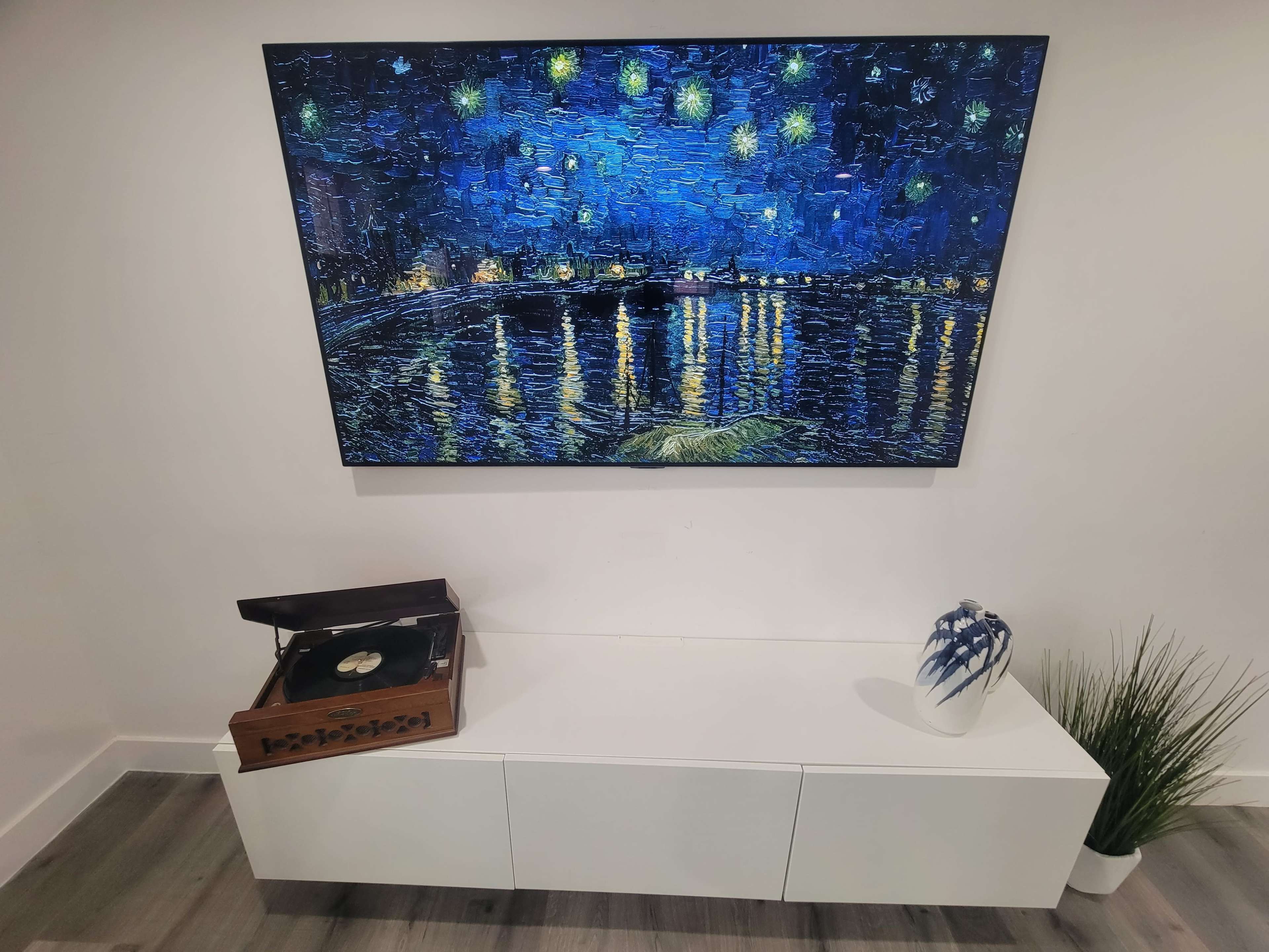 A modern room with a large painting of a night scene displayed on the wall, a record player on a white cabinet, and a decorative vase on the surface.
