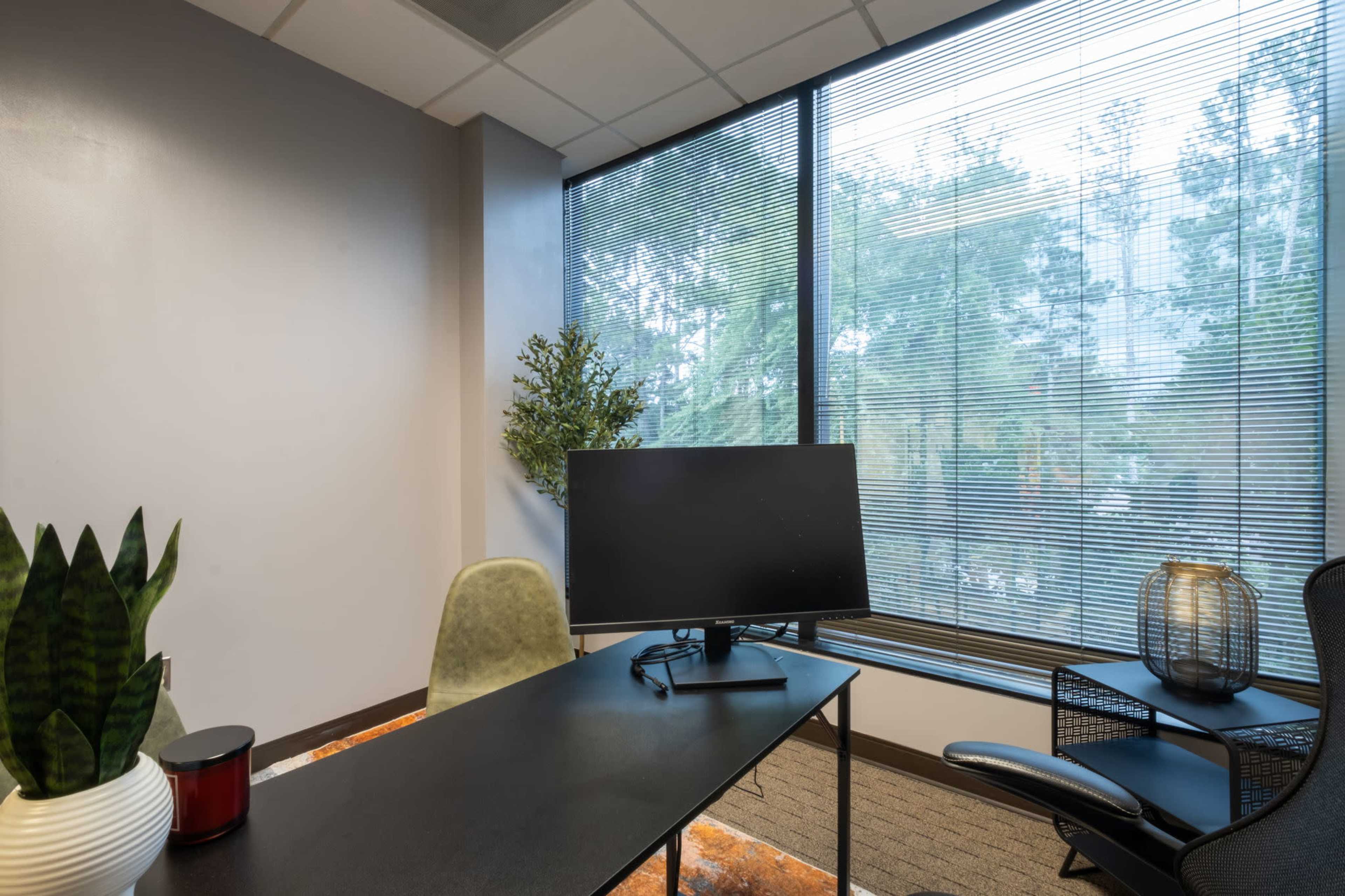 The image shows a modern office space with a desk, a computer monitor, a chair, and large windows covered with blinds.