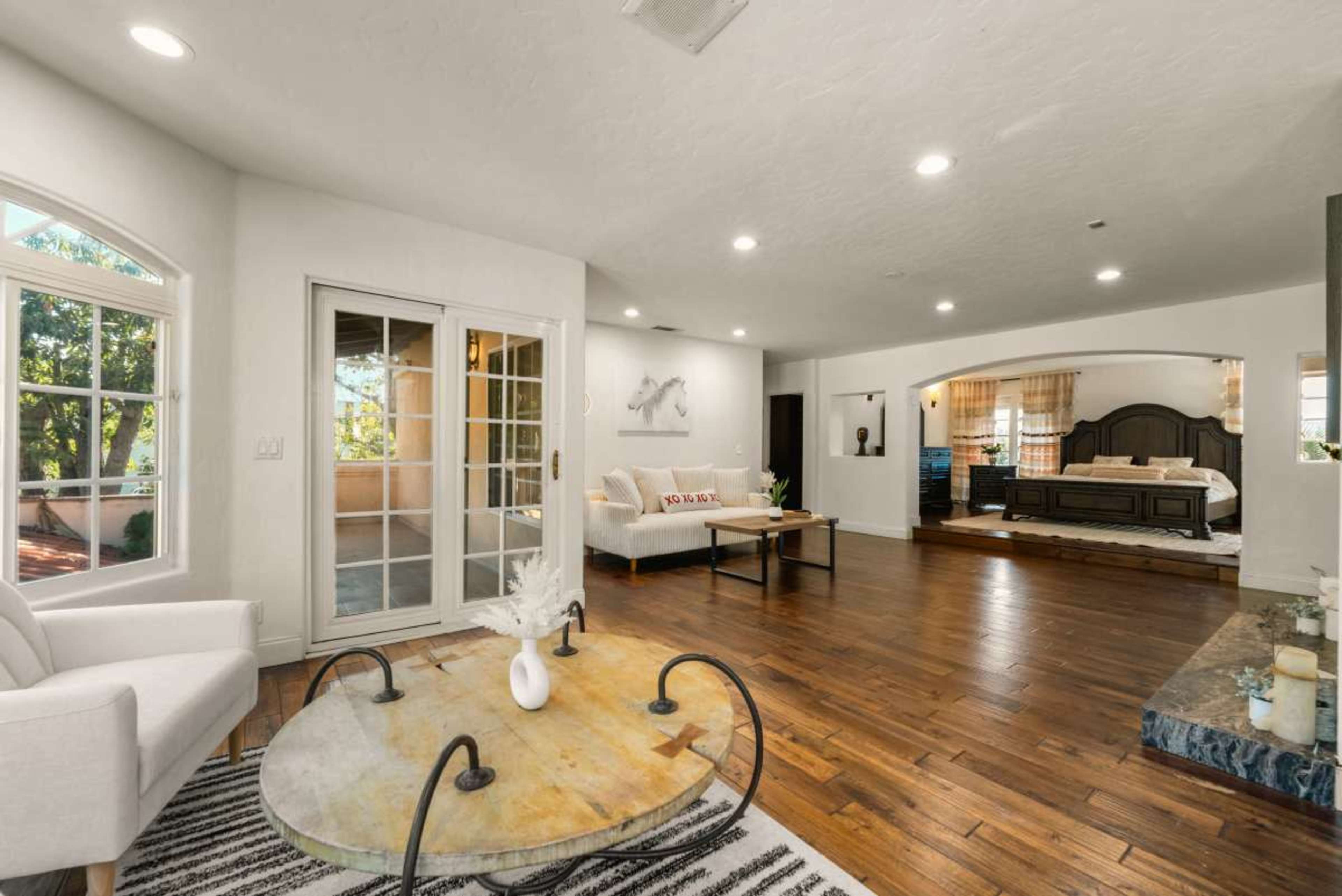 The image shows a spacious living area with hardwood floors, a round coffee table, and access to a patio through French doors.
