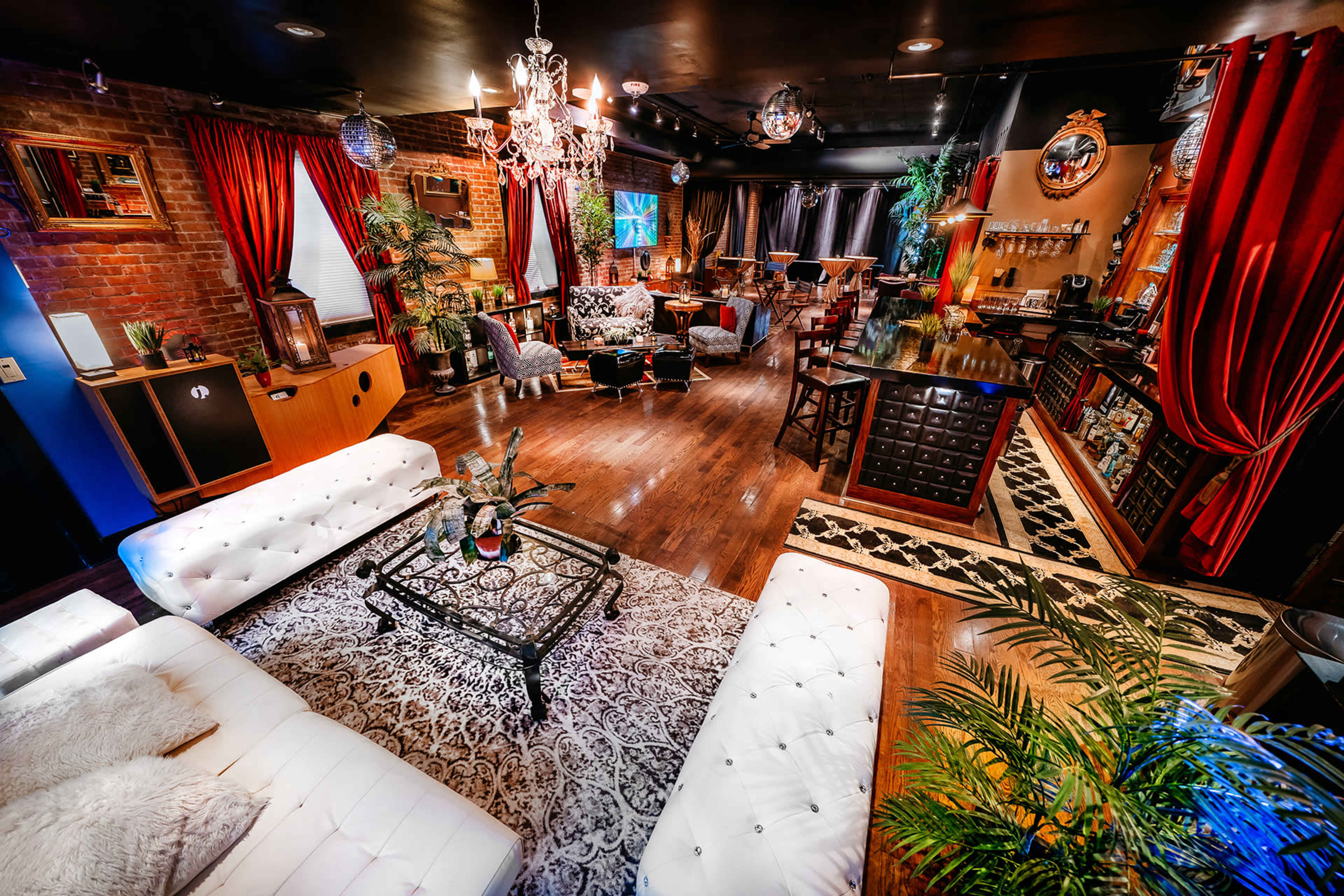 A stylish lounge area filled with plush seating, decorative plants, and a bar, illuminated by chandeliers and accent lighting.