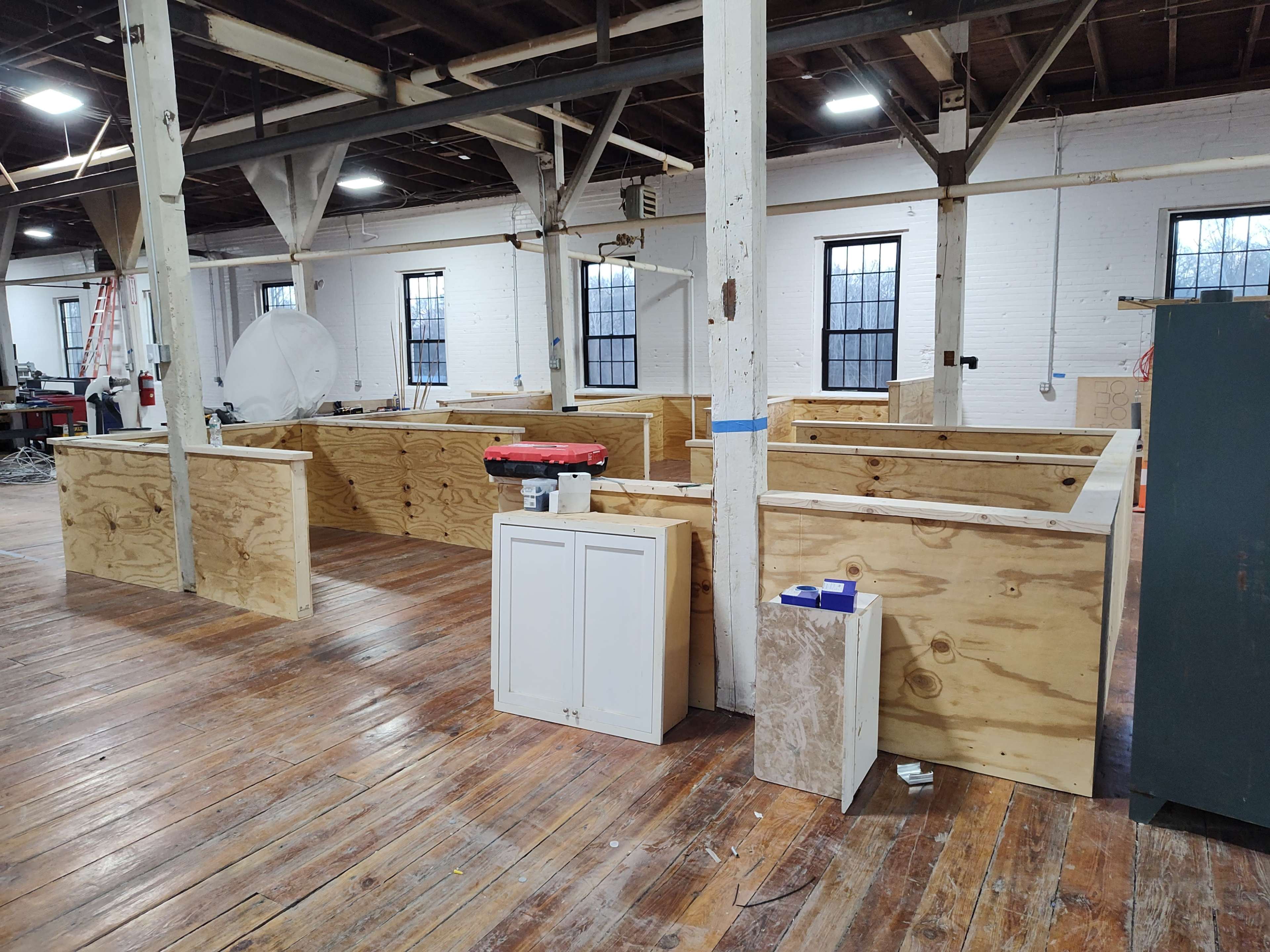 The image shows an unfinished interior space with wooden structures, including workstations and storage units, on a hardwood floor.