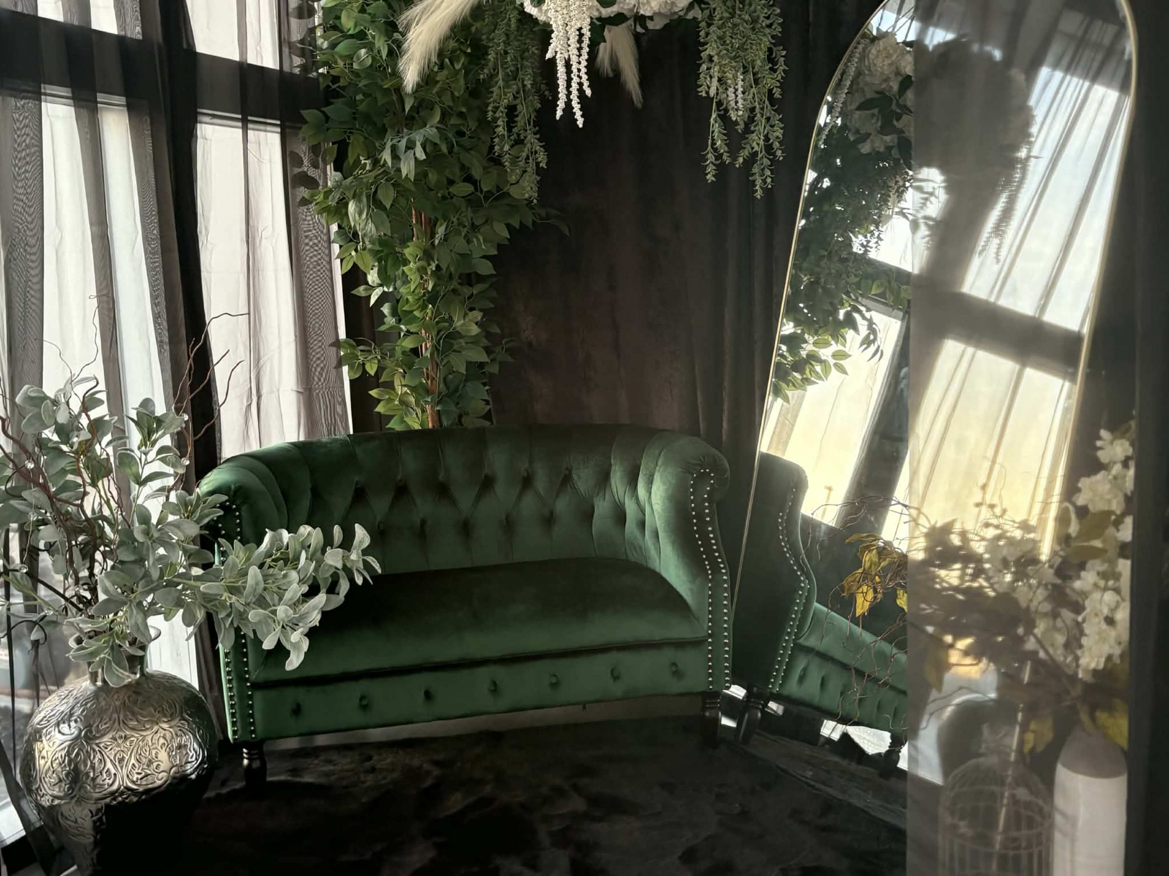 The image features a green velvet tufted sofa positioned near a large mirror, surrounded by greenery and decorative plants.