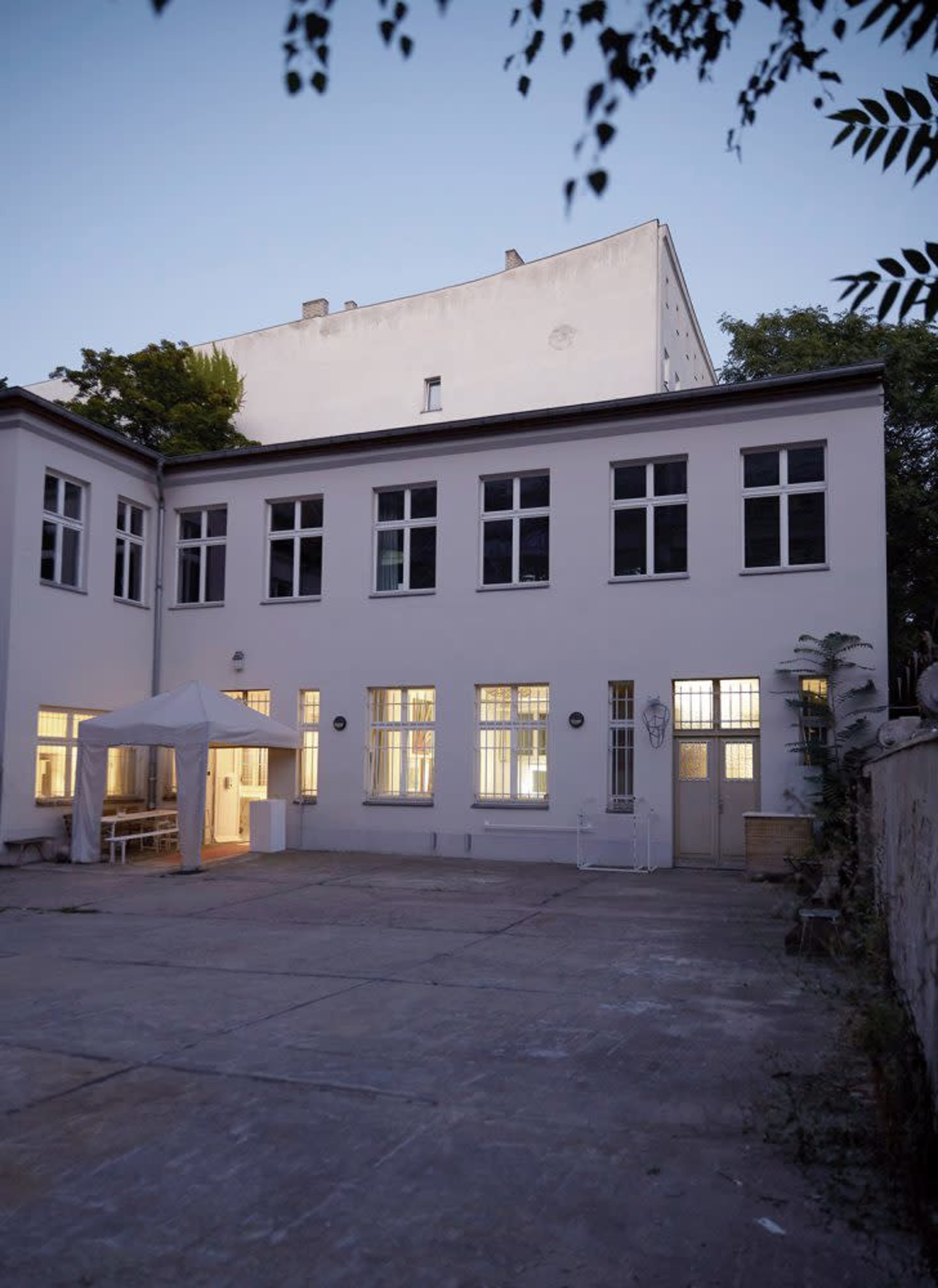 A white building with multiple windows and a tent set up in a paved courtyard during twilight.