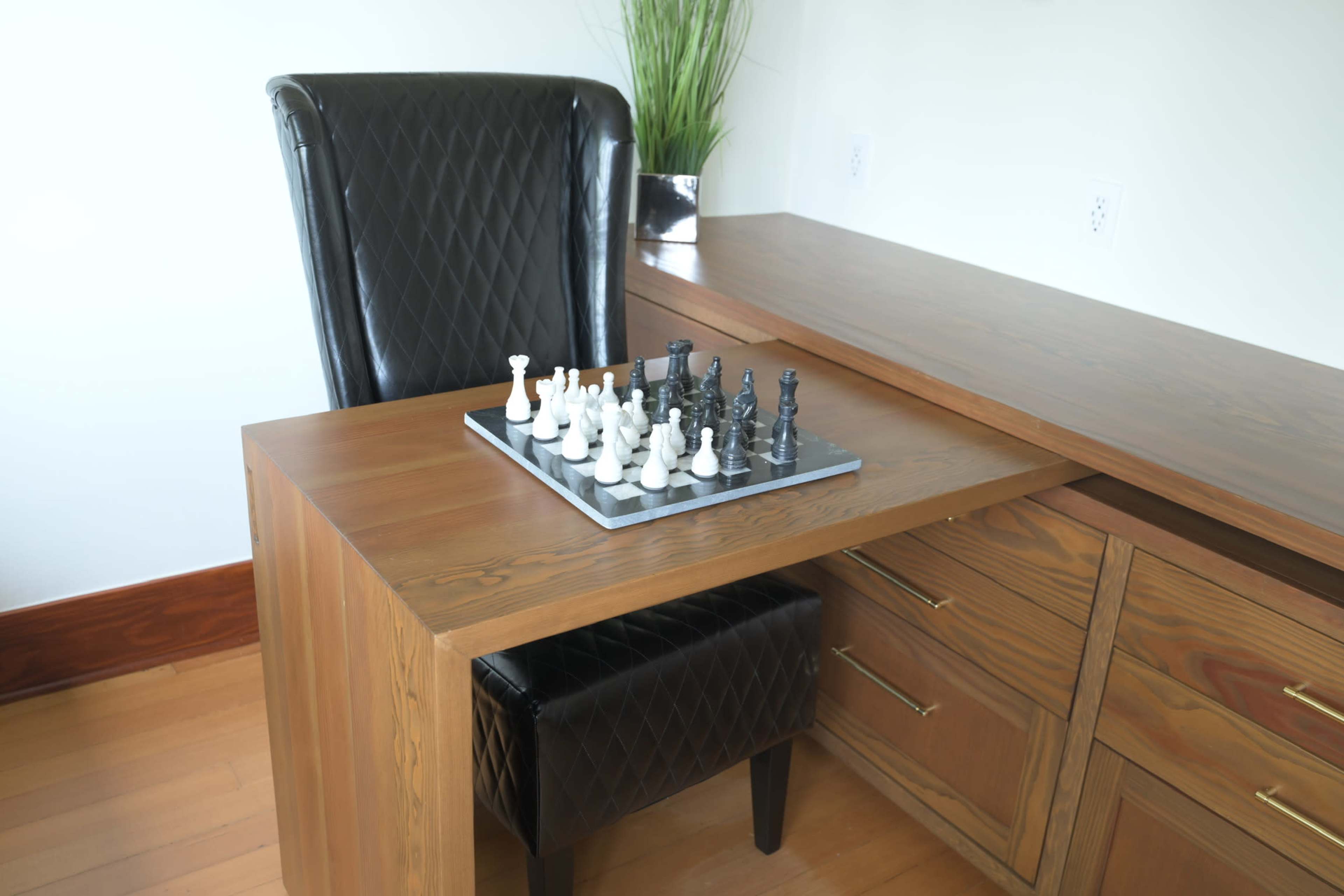 A chessboard with white and black pieces is placed on a wooden desk next to a black upholstered chair in a modern workspace.