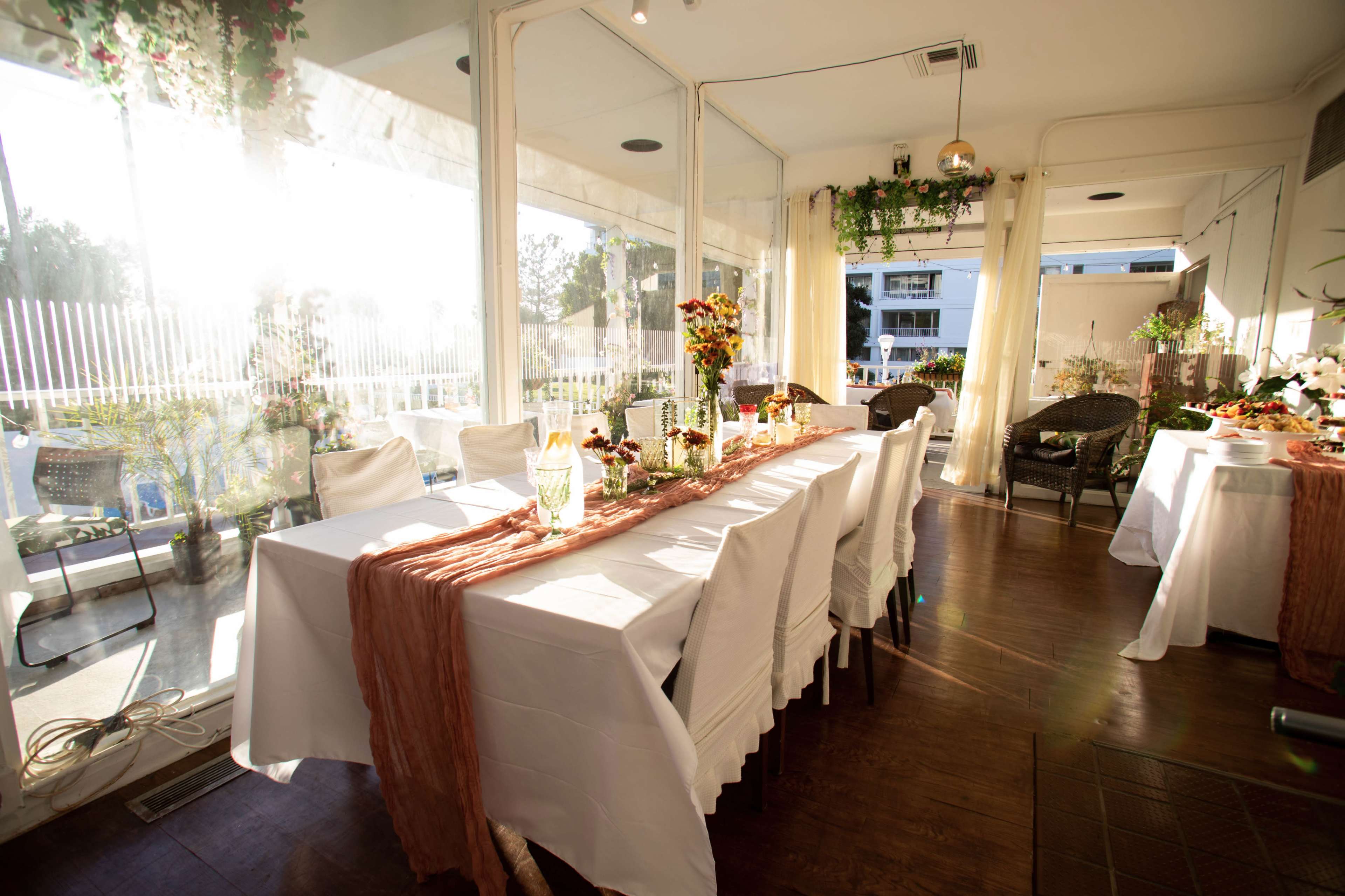 A well-set dining area with white tablecloths, elegant floral centerpieces, and sunlight streaming in through large glass windows.