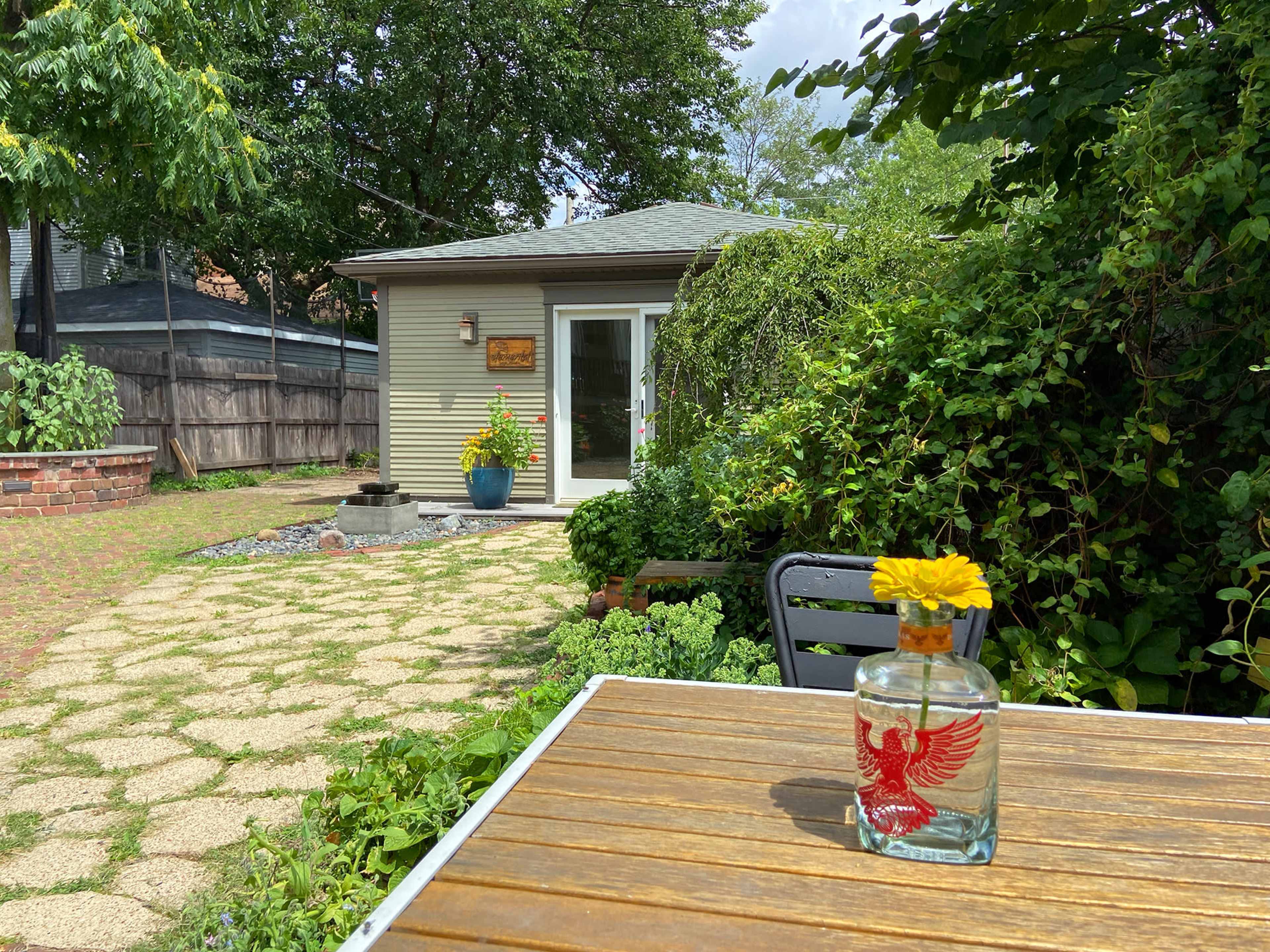 A small garden space features a wooden table with a decorative bottle and a flower, while a house and lush greenery are in the background.