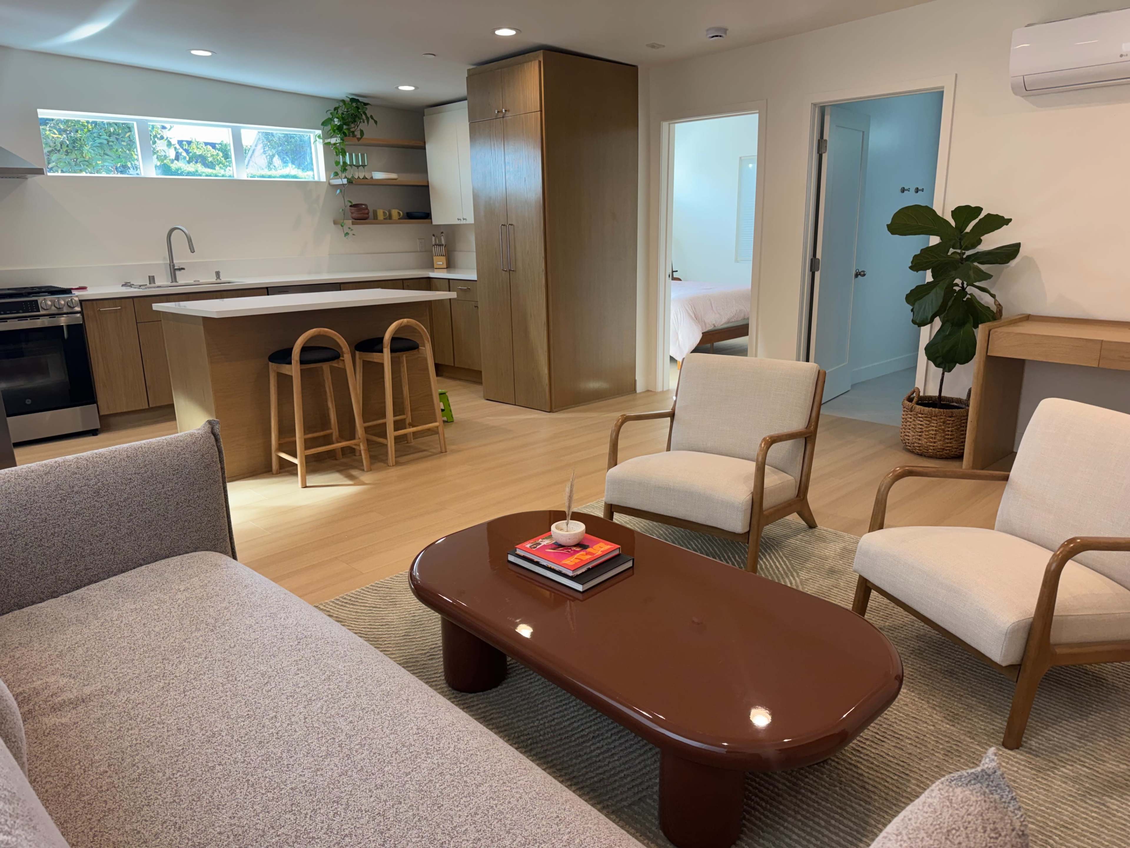 The image shows a modern living space featuring a sofa, two armchairs, a coffee table, and an open kitchen area with wooden cabinetry and bar stools.