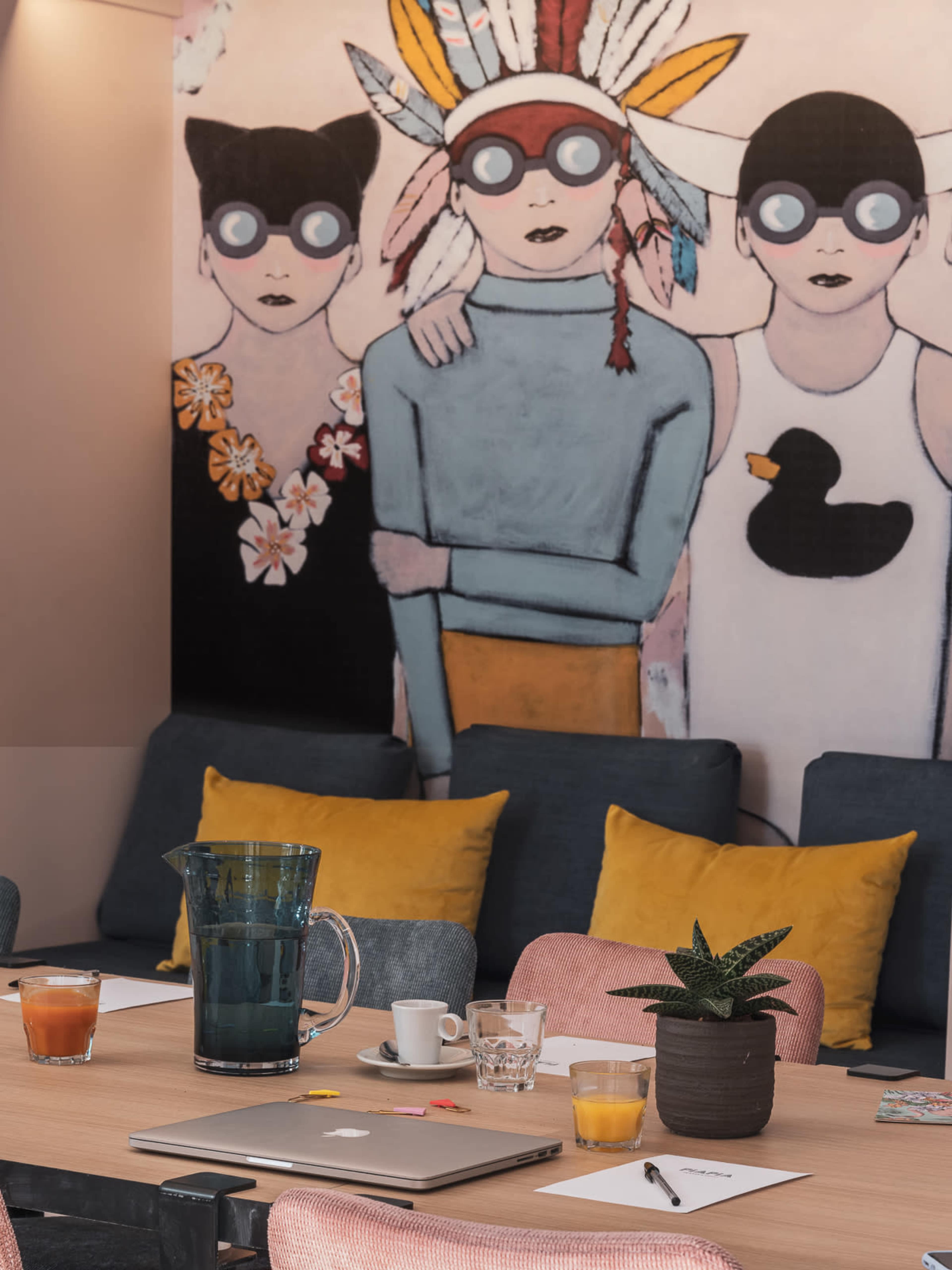 The image shows a modern meeting room with a table covered in notebooks, drinks, and a laptop, alongside a colorful mural of three figures with distinctive headgear.