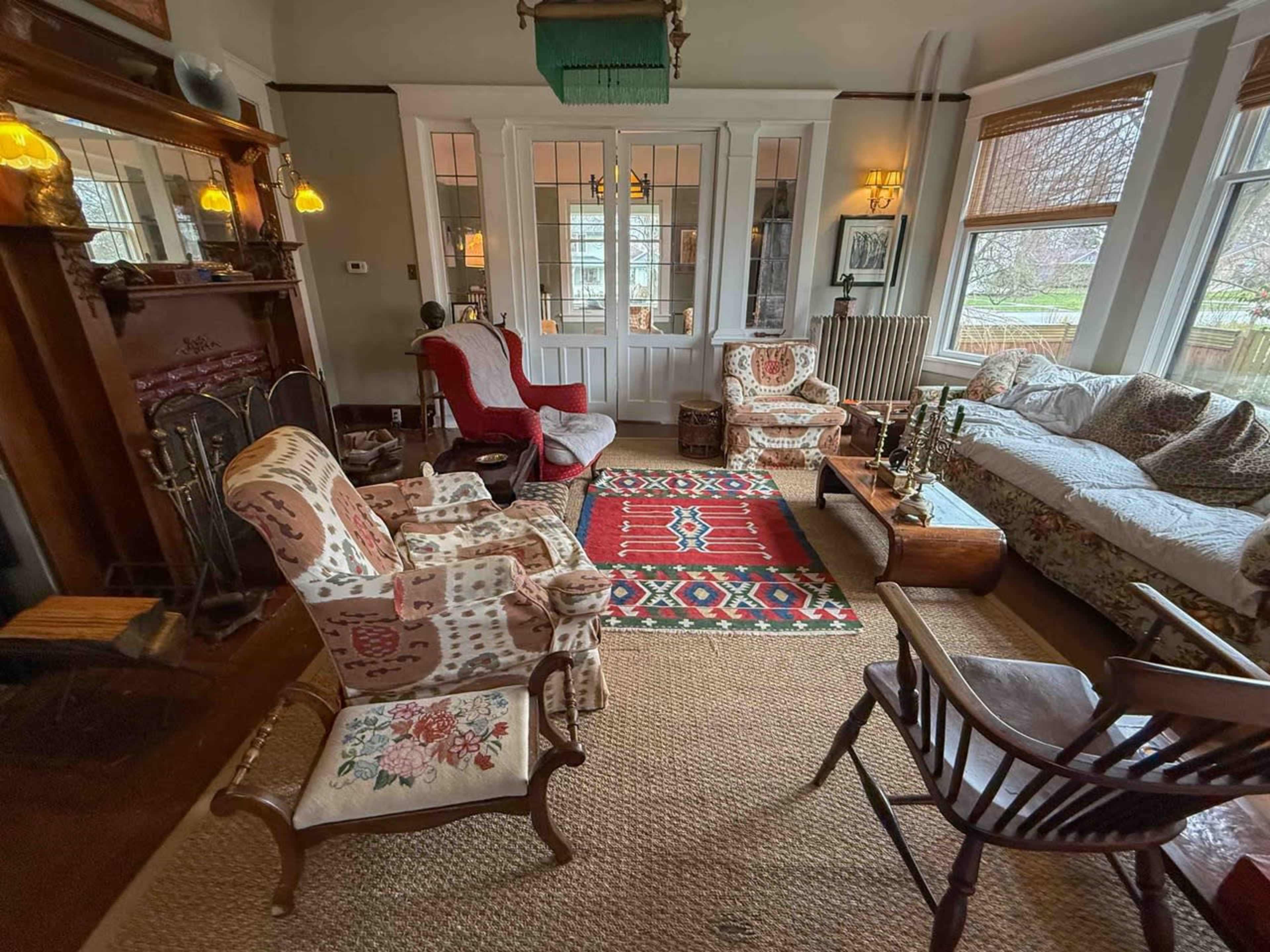 The room features a mix of patterned seating, a patterned rug, and wooden furniture arranged around a central area, with large windows allowing natural light to enter.
