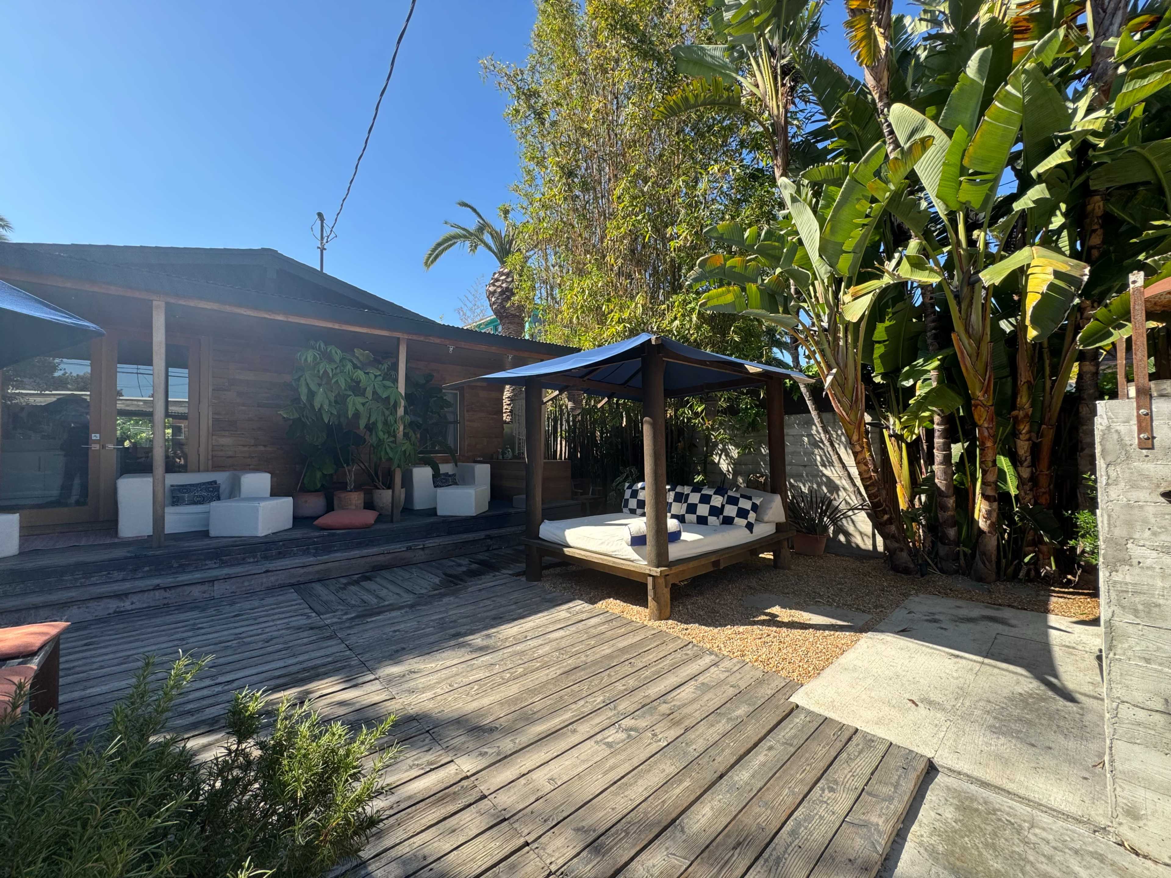 The image shows a sunlit outdoor space with a raised wooden deck, a lounge area featuring a round bed under a canopy, surrounded by tropical plants and a stone pathway.
