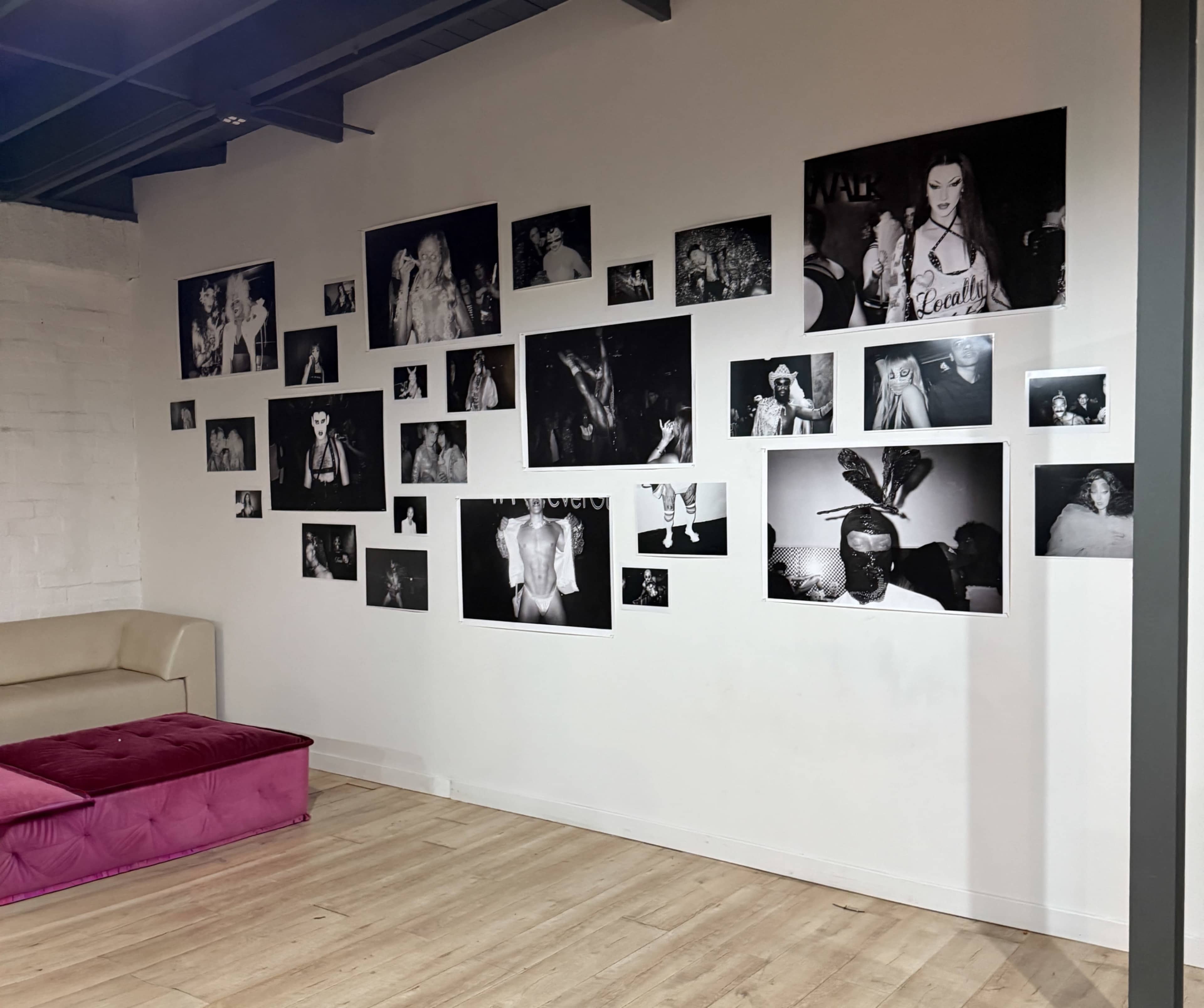The image features a wall adorned with numerous black-and-white photographs, showcasing a variety of people and scenes, accompanied by a pink velvet couch in the foreground.