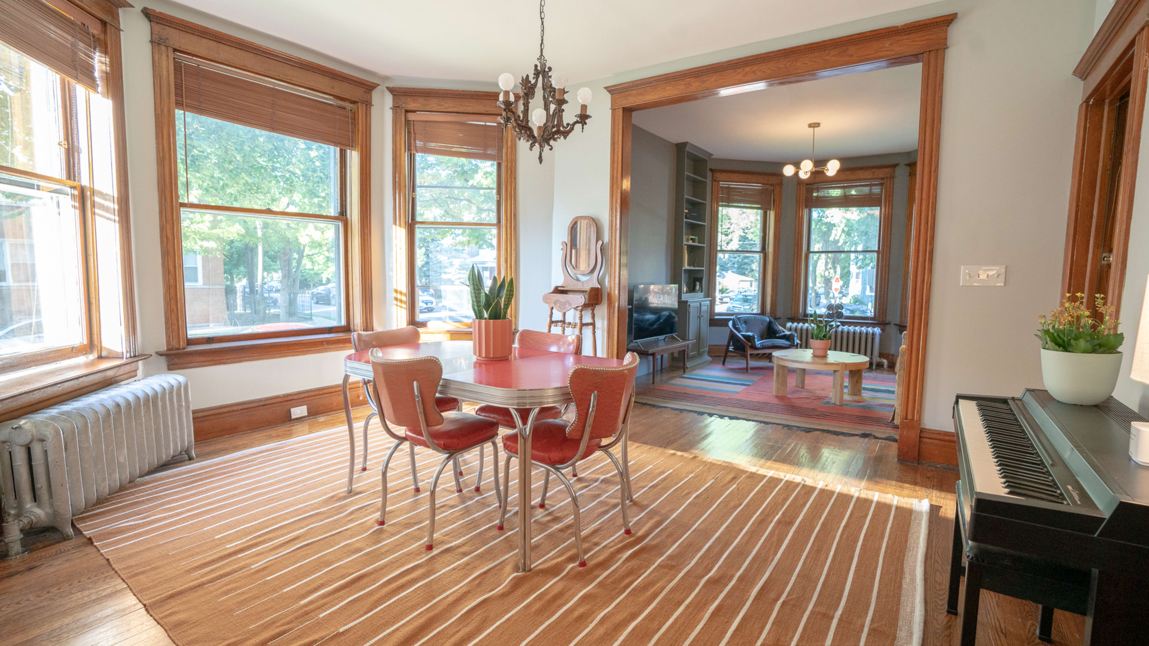 A dining area with a round table and four chairs is situated next to large windows, leading into a living room with a piano and seating arrangement.