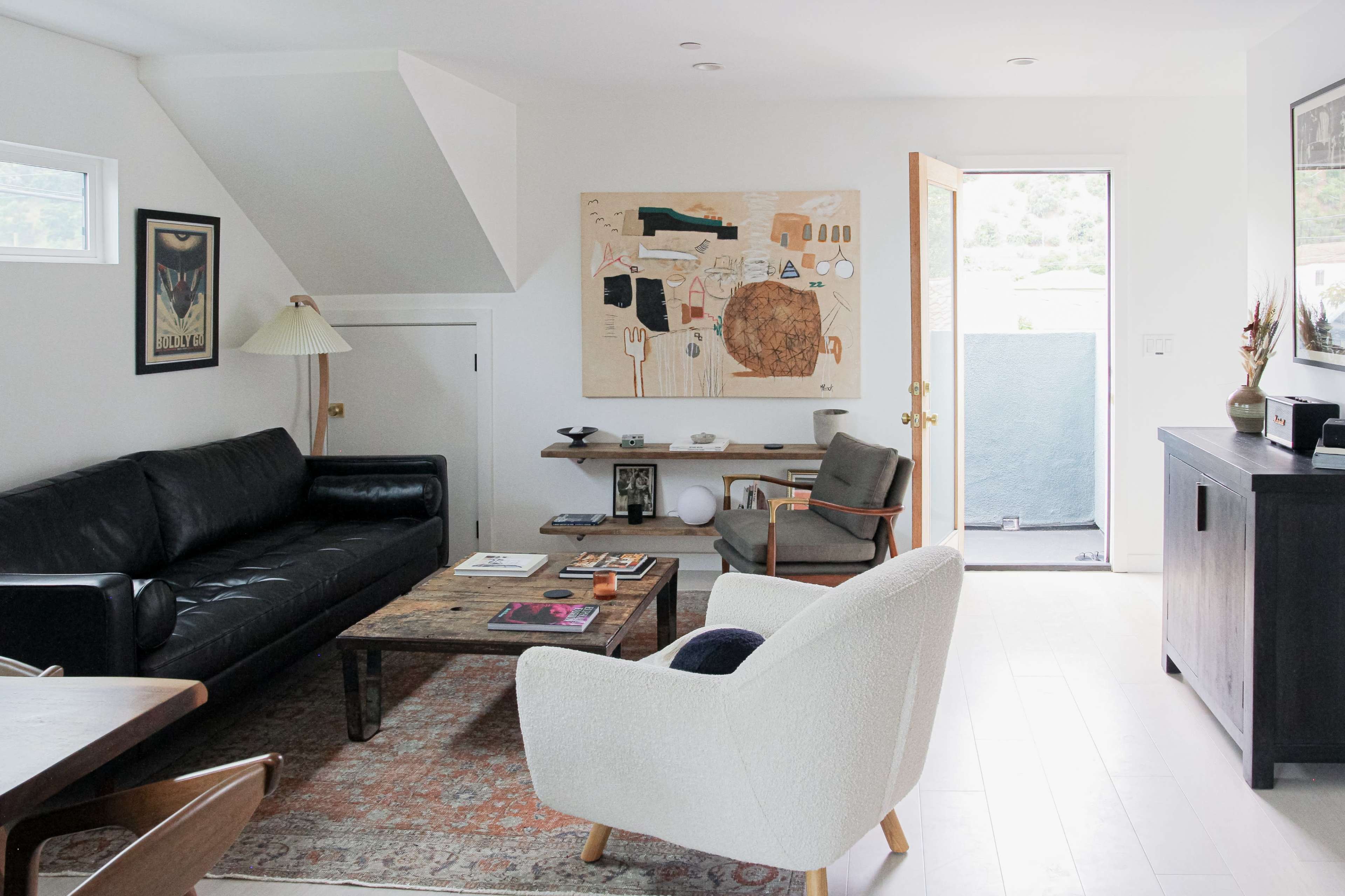A minimalist living room features a black sofa, a wooden coffee table with books, and an open door leading to an outdoor area.