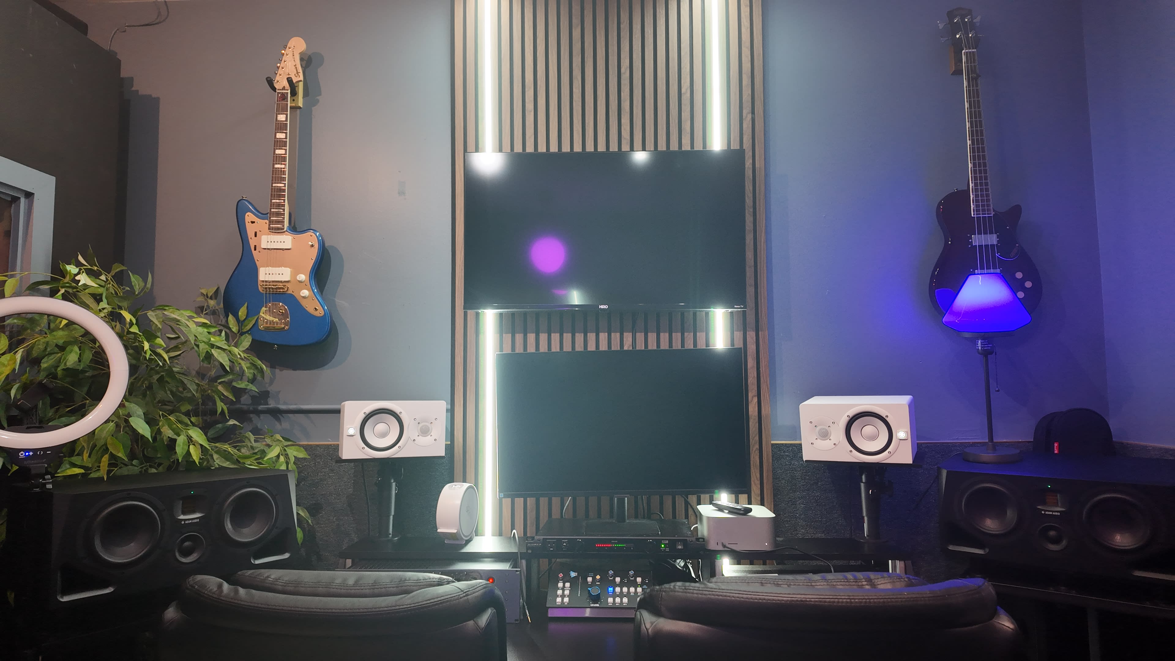 The image shows a modern music production studio featuring two guitars mounted on the wall, a dual-screen setup in the center, and various audio equipment on a desk.