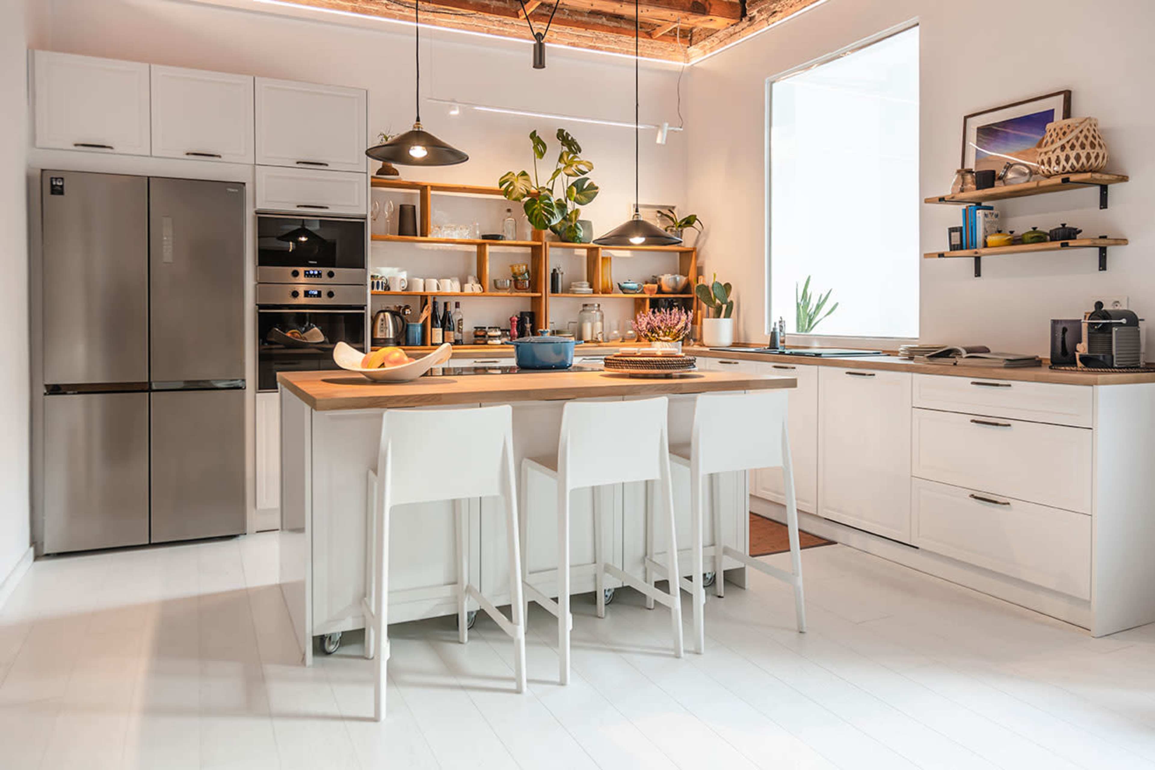 A modern kitchen featuring a gray refrigerator, white cabinetry, a wooden island with seating, and shelves displaying plants and kitchenware.