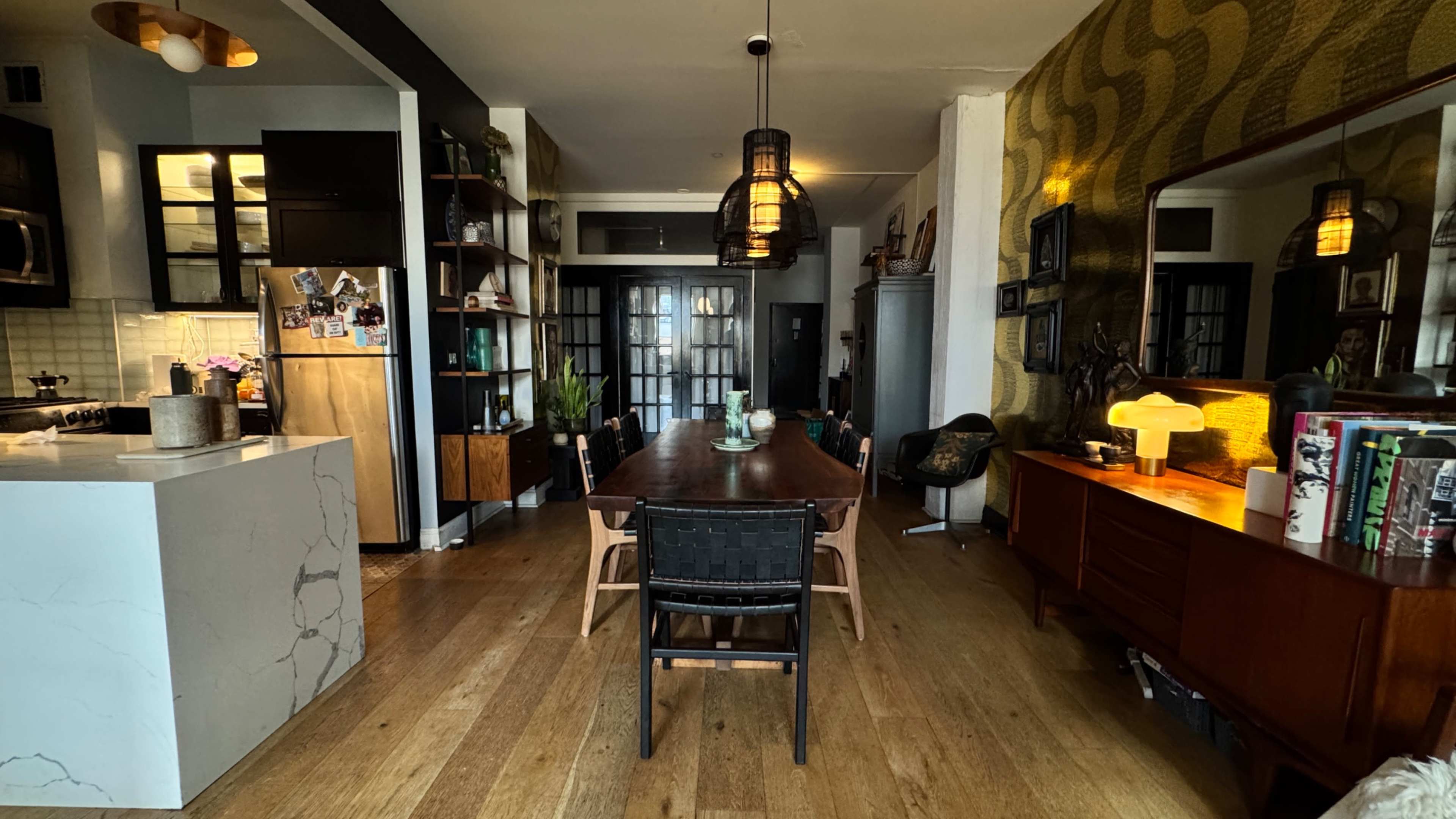 The image shows a modern dining area featuring a long wooden table surrounded by black and wooden chairs, with an open kitchen and various decorative elements in the background.