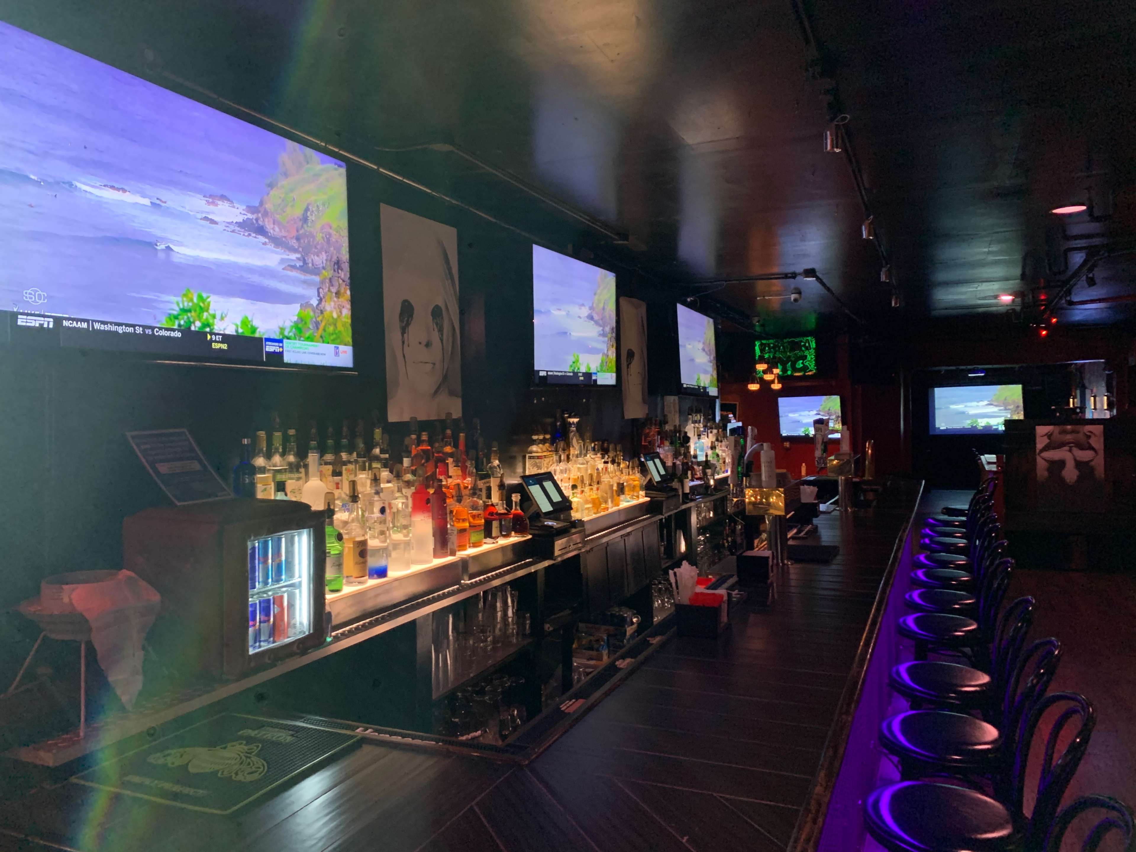 A dimly lit bar with multiple televisions displaying ocean scenery, a well-stocked liquor shelf, and rows of bar stools.