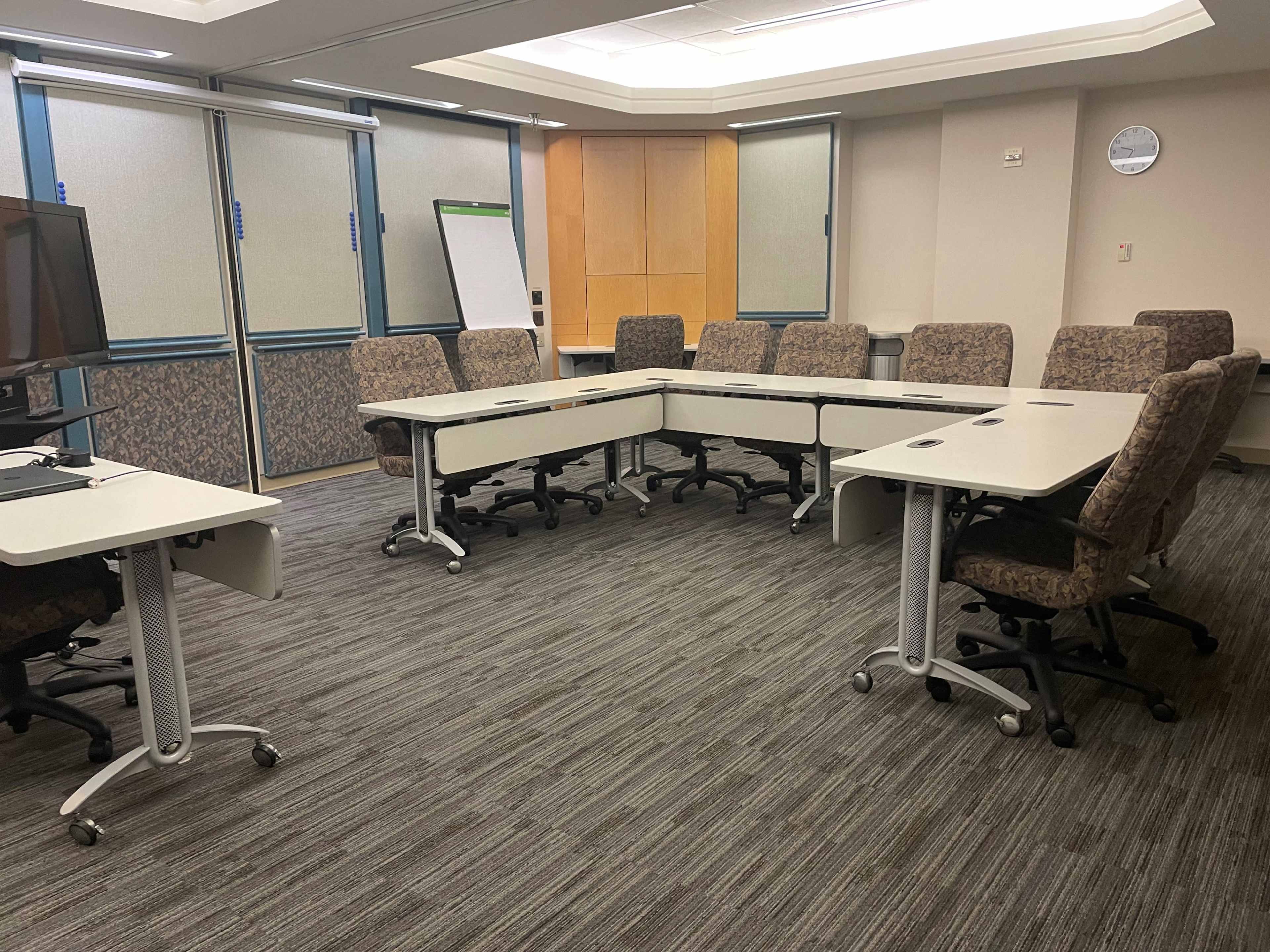 A conference room is set up with a large, oval table surrounded by eight chairs, with a television and a whiteboard positioned at one end.