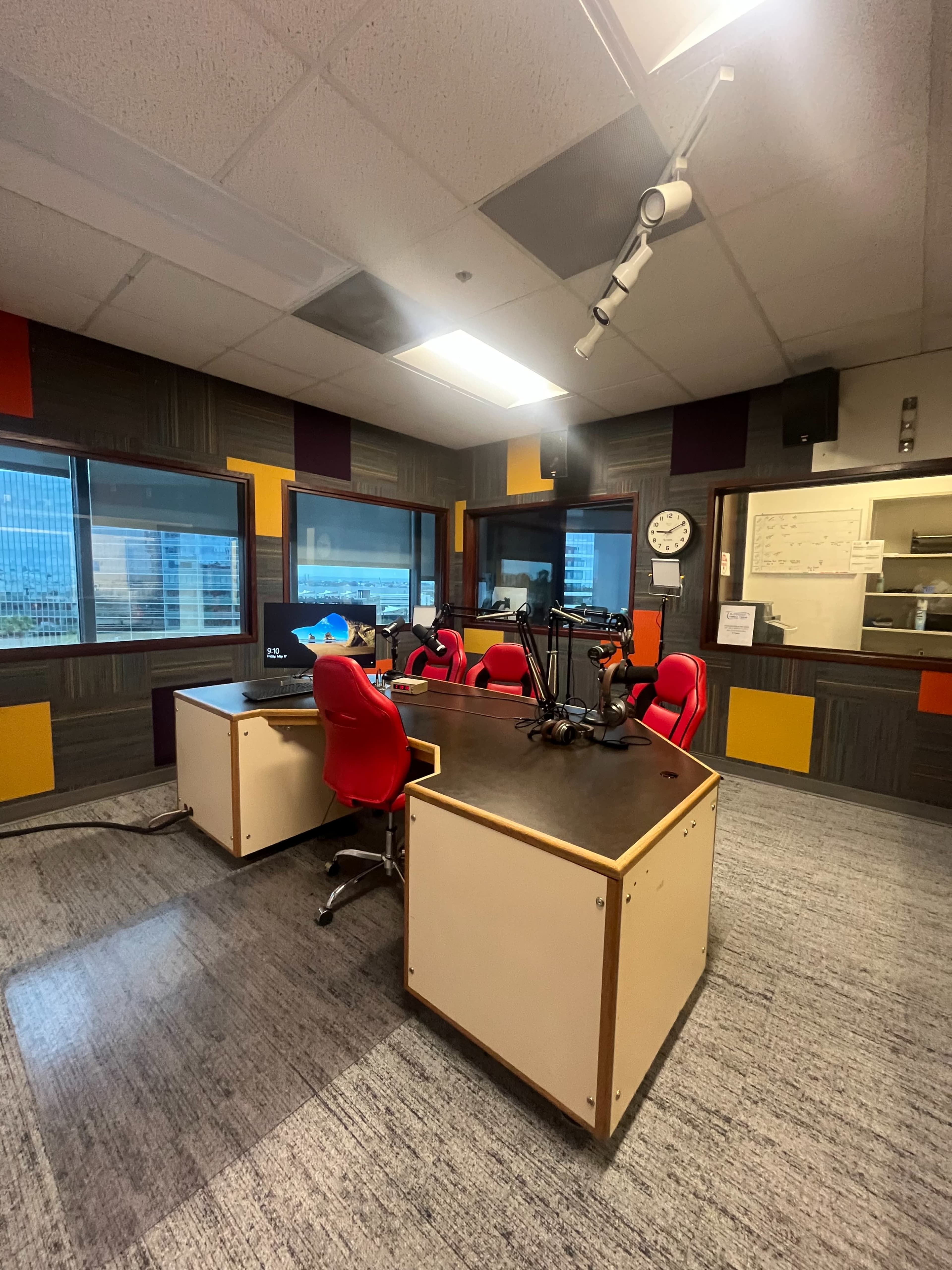 A radio studio with two red chairs, a large desk, and equipment set up against a wall of windows.