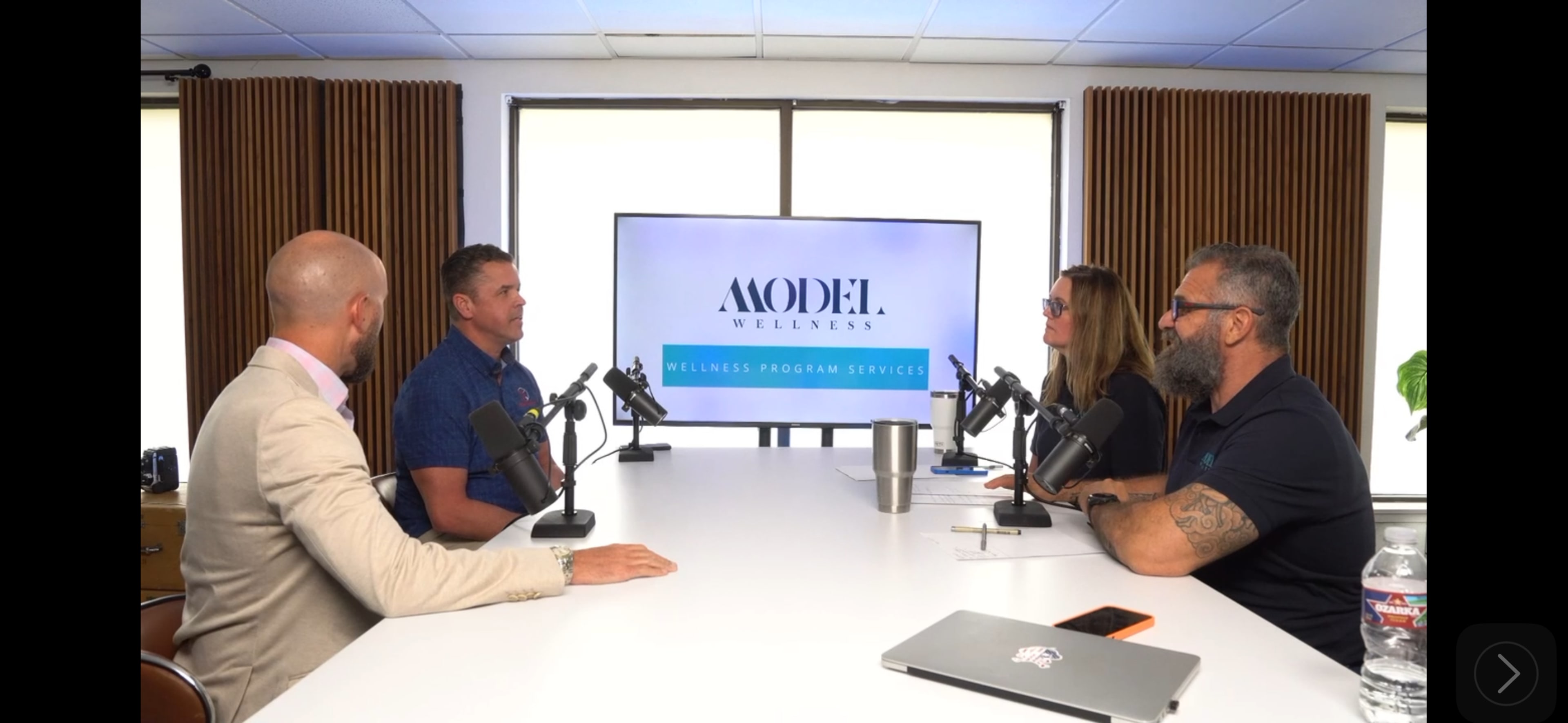 A group of four individuals is seated around a conference table, engaged in a discussion while a large screen displays the logo and name of a wellness program.