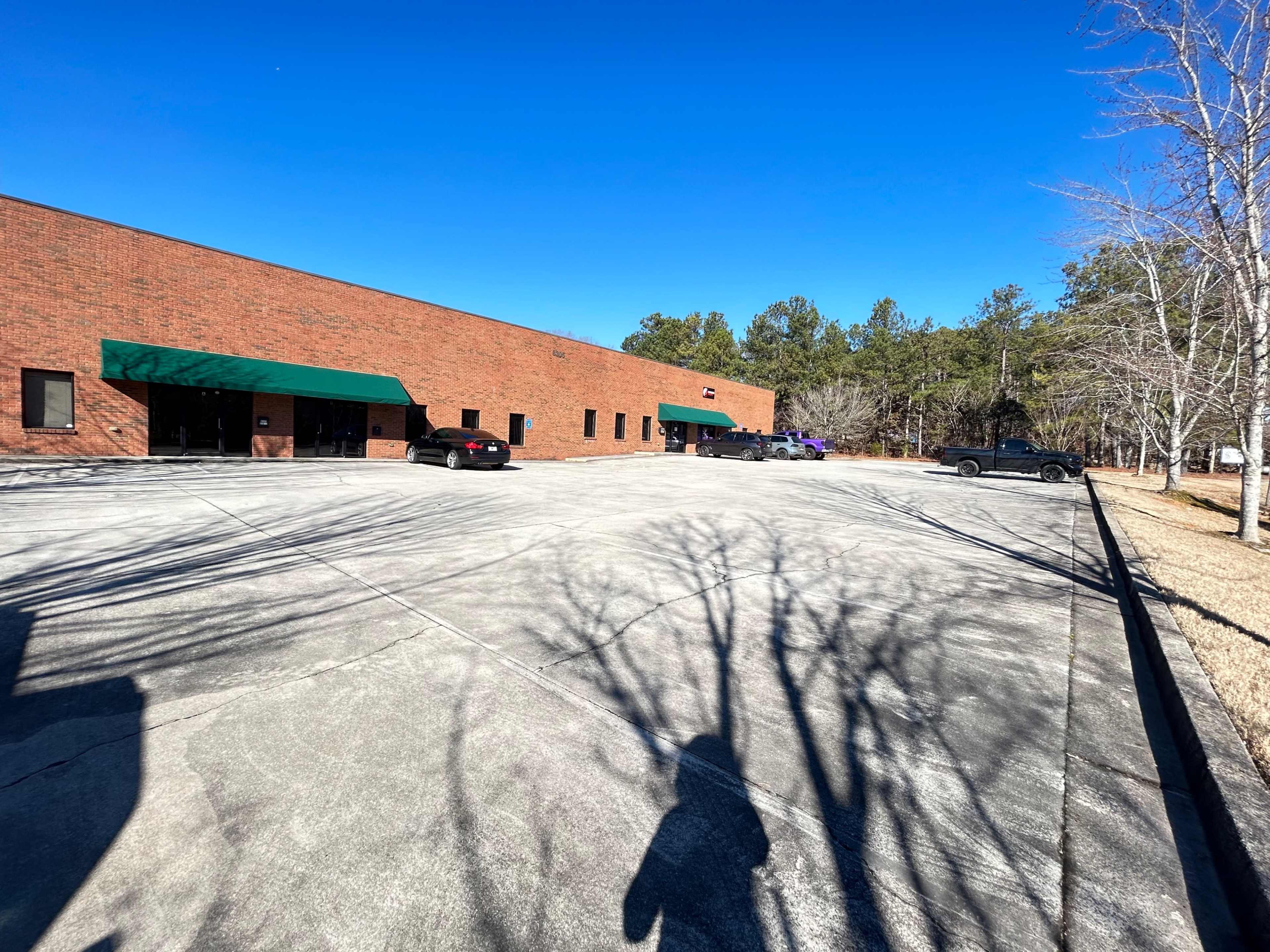 The image shows a parking lot in front of a brown brick building, partially shaded by trees, with several parked vehicles.