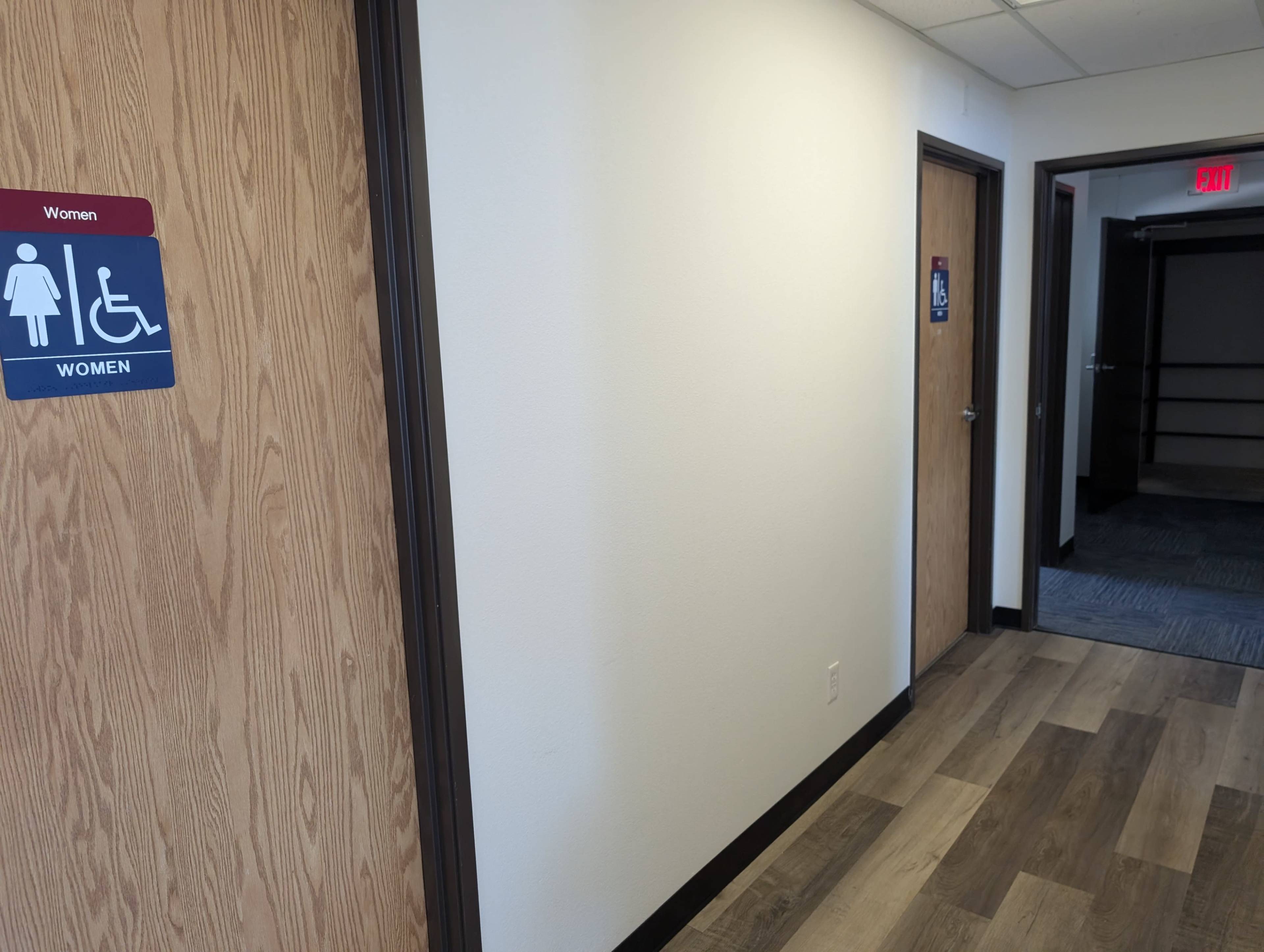 The image shows a corridor with a sign for women's restrooms on the left and a door leading to another area on the right.