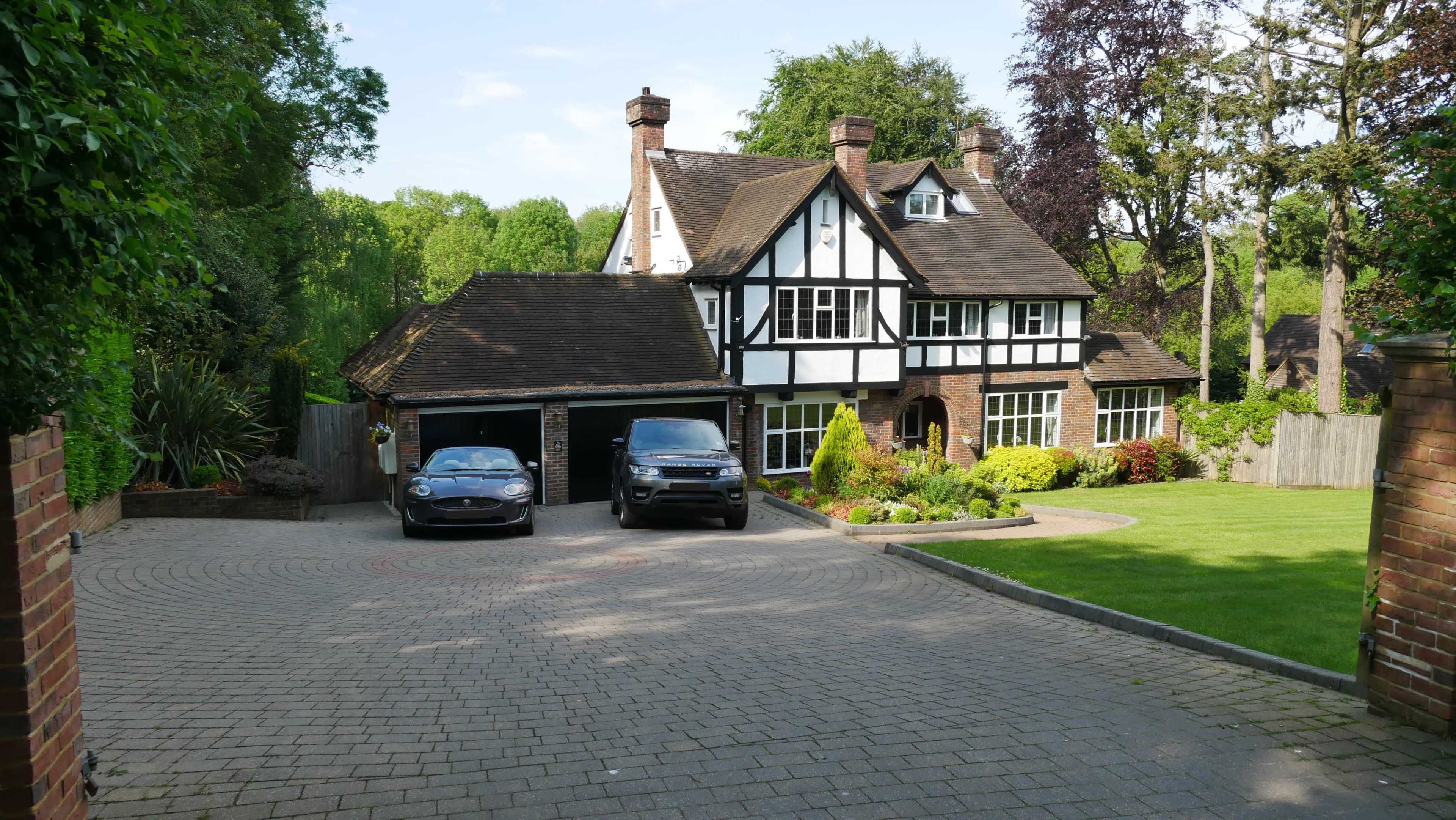 A large, two-story house with a black and white Tudor-style facade, two cars parked in the driveway, and a well-maintained garden surrounding the property.