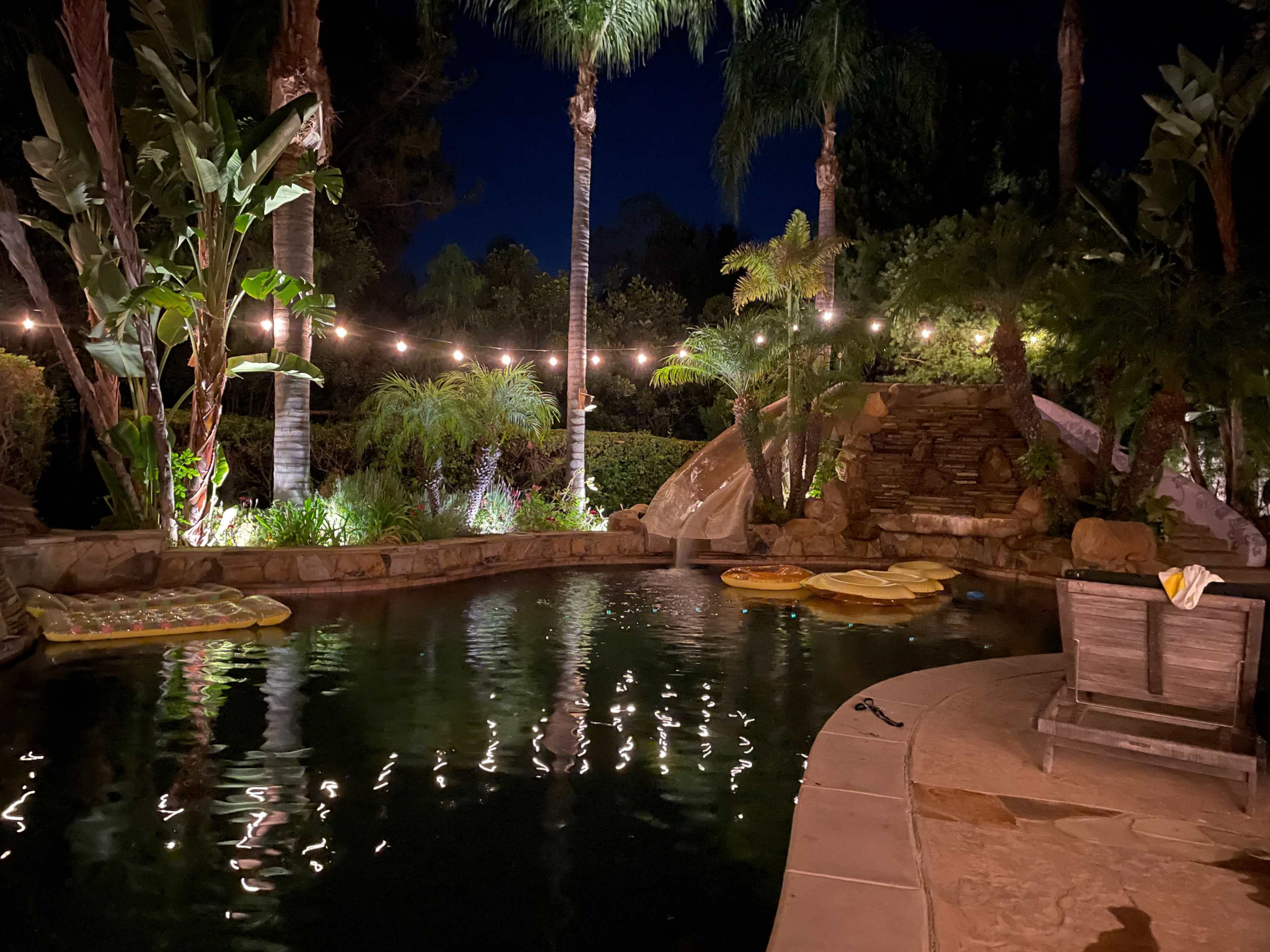 The image shows a well-lit backyard pool area surrounded by palm trees and lush greenery, with floating pool toys and string lights creating a cozy atmosphere at night.